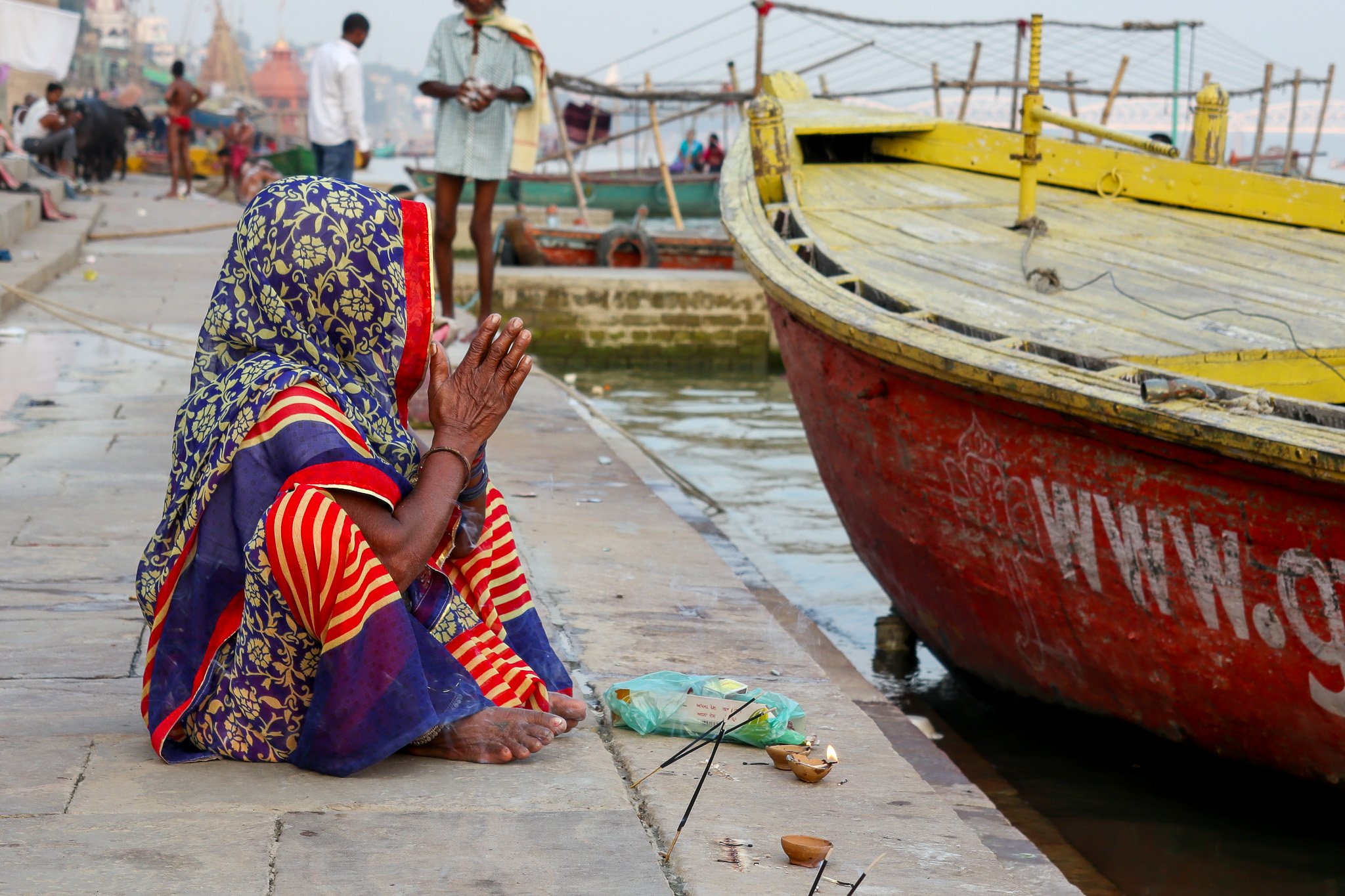A woman in colorful traditional attire performing a prayer or ritual near a riverbank with boats in the background in India.
