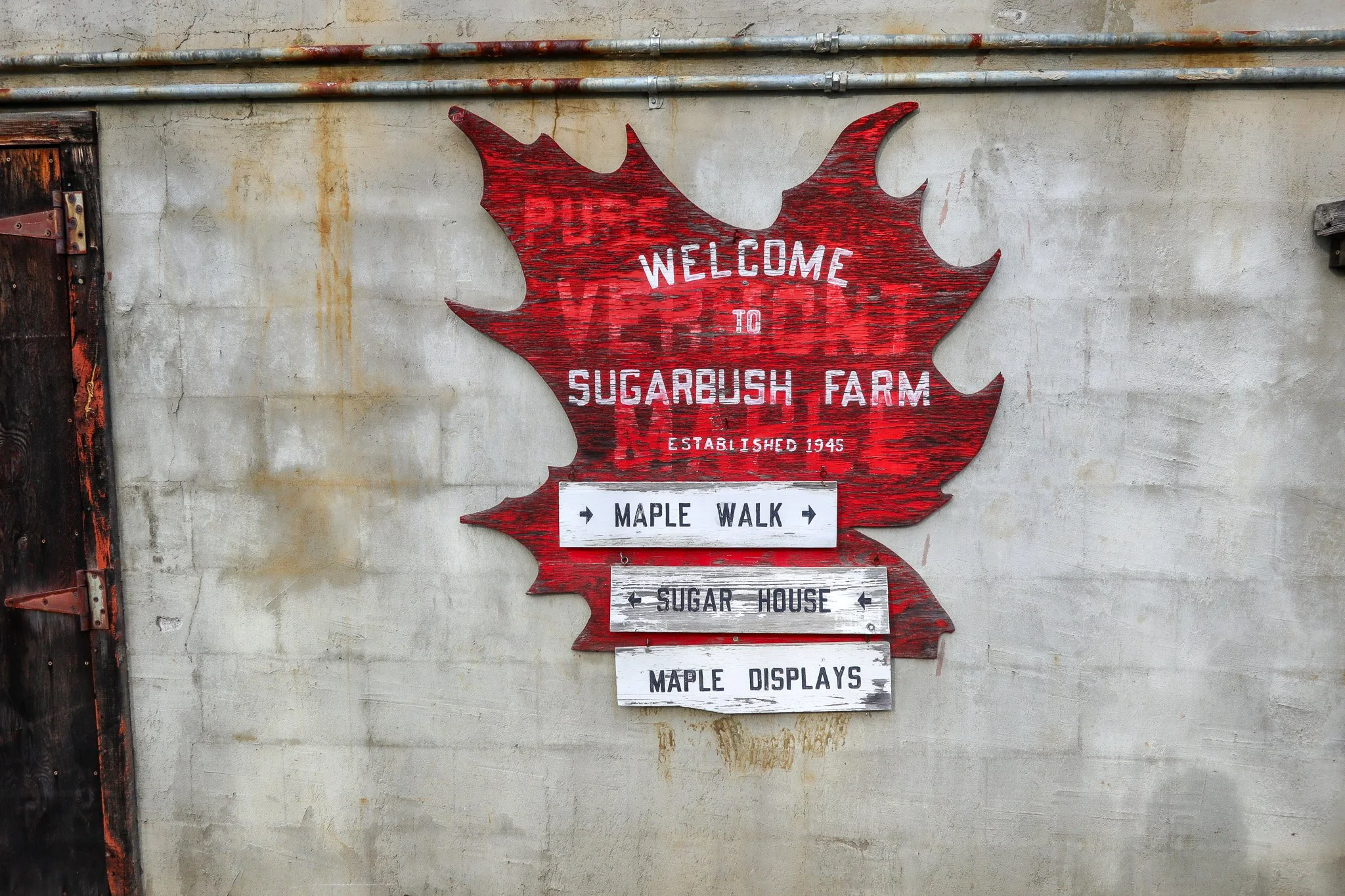 Red wooden sign shaped like a maple leaf with white text welcoming visitors to Sugarbush Farm, established in 1945. Below are three white directional signs with black text pointing to Maple Walk, Sugar House, and Maple Displays, mounted on a gray con