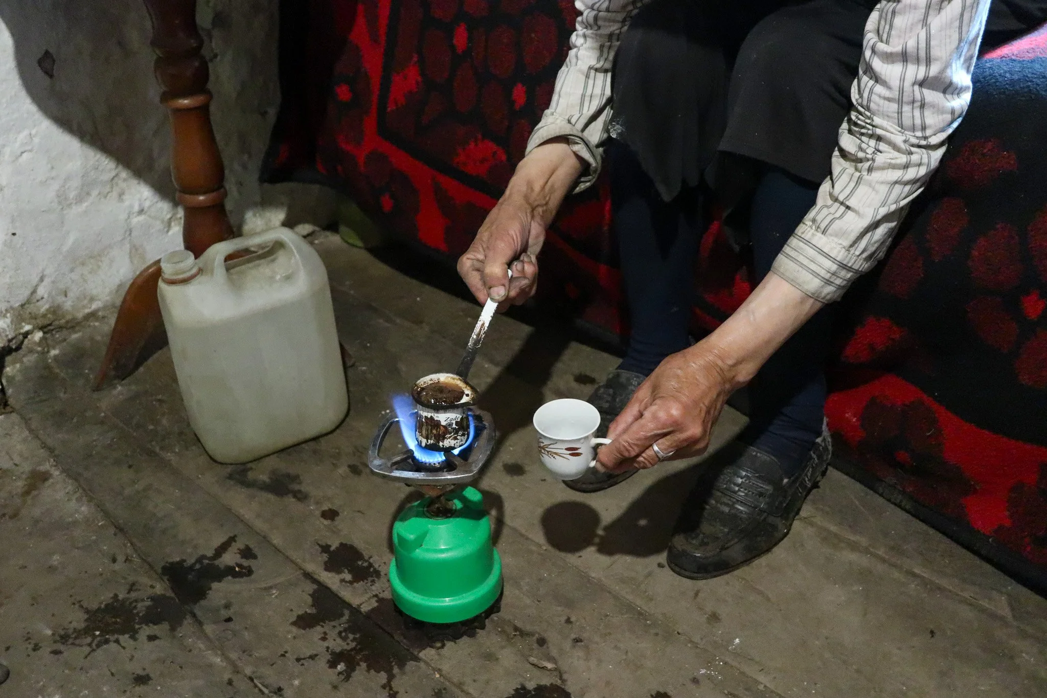 An elderly person is heating coffee on a small portable stove, holding a cup in one hand and using a spoon to stir. On the ground, there is a gallon water container. The scene appears to be indoors with a wooden floor and a patterned rug, with part o