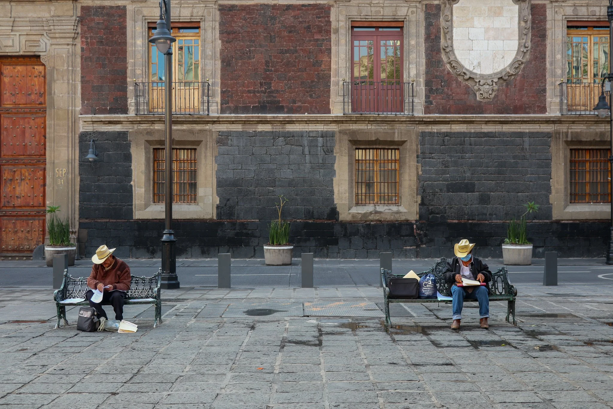 Two people wearing hats and face masks sit on separate benches in a city square, reading or writing. There are bags and books with each person, set against a backdrop of a historic building with barred windows and decorative stonework.