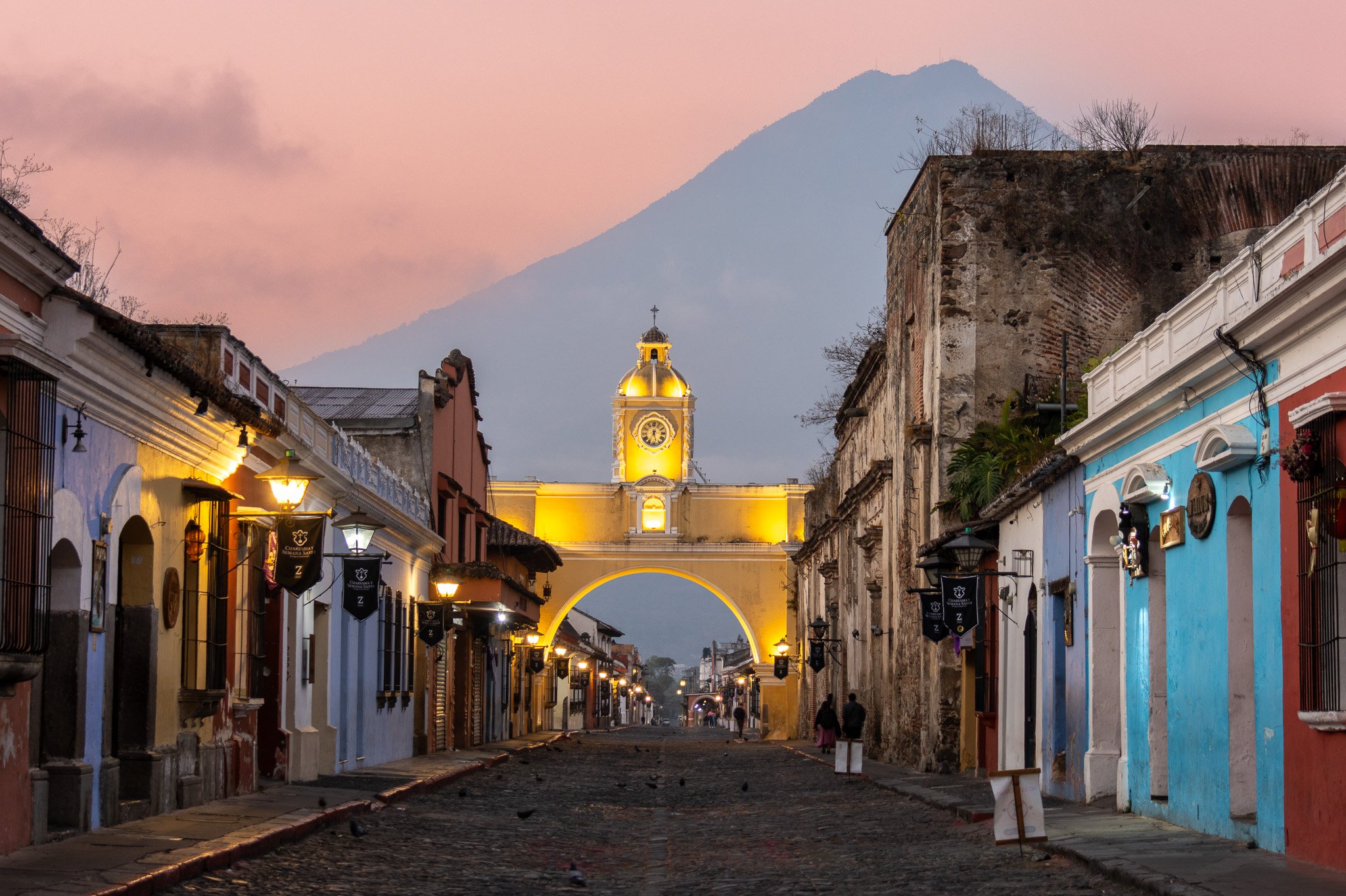 Colorful street with colonial architecture and old lamp posts leading to a yellow clock tower with a dome, set against a mountain at sunset.