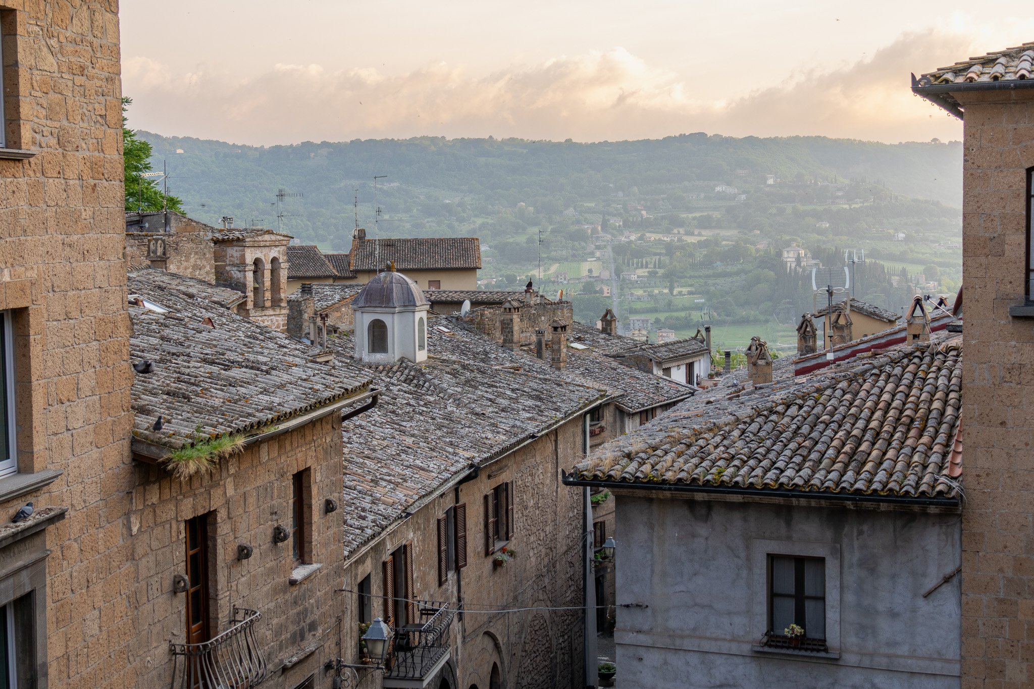 View of an Italian hillside town with rustic stone buildings and tiled roofs, overlooking rolling green hills in the distance during sunrise or sunset.