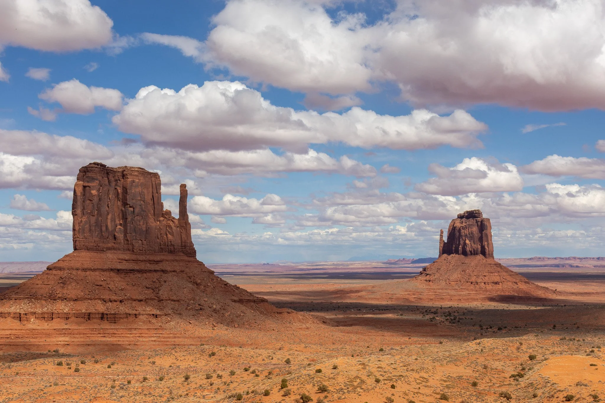 Two large sandstone buttes in a desert landscape under a partly cloudy sky, with sparse vegetation on the ground.