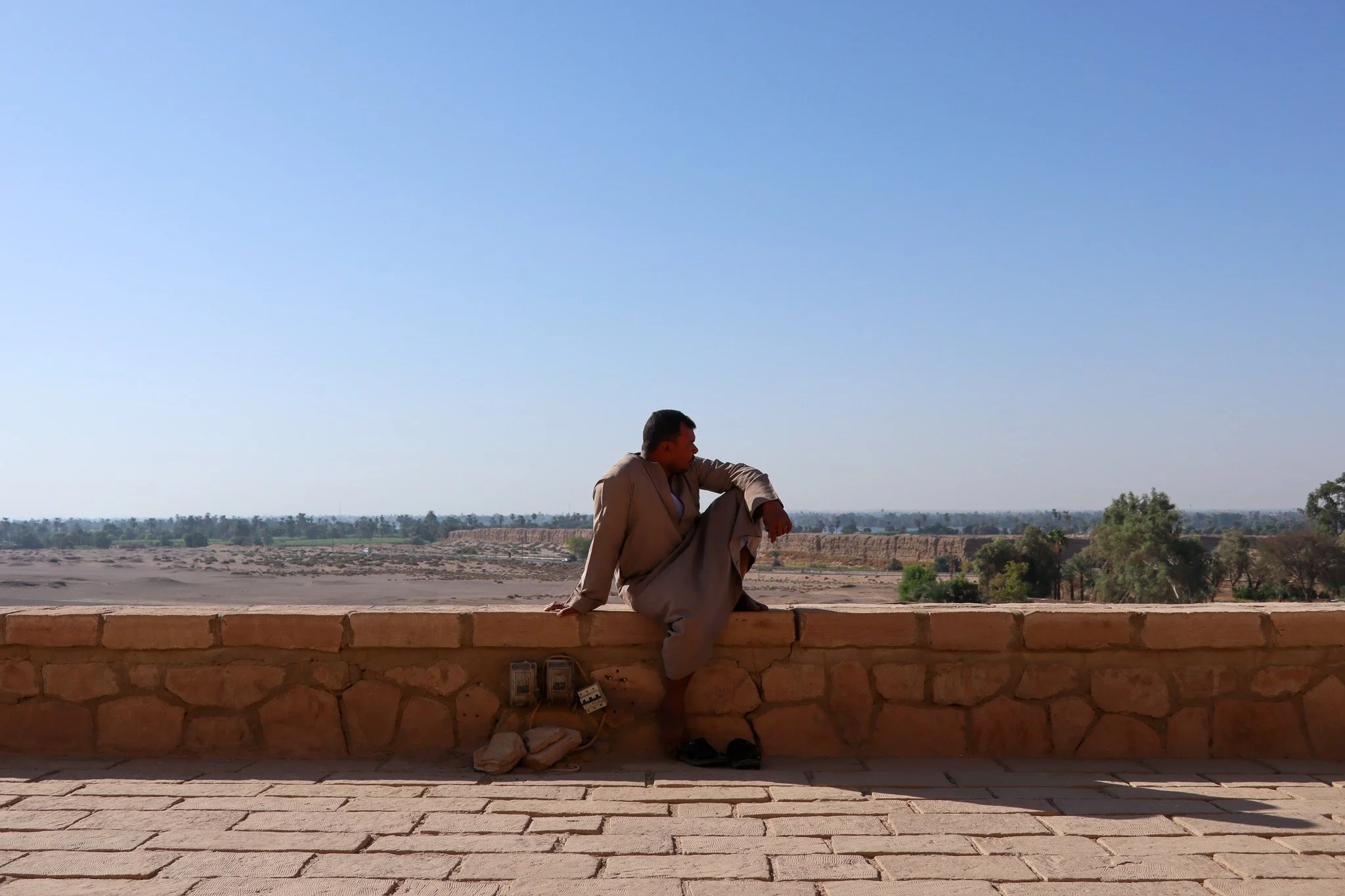 A man dressed in traditional clothing sitting on a stone wall, looking out over a barren landscape with sparse trees and a clear blue sky.