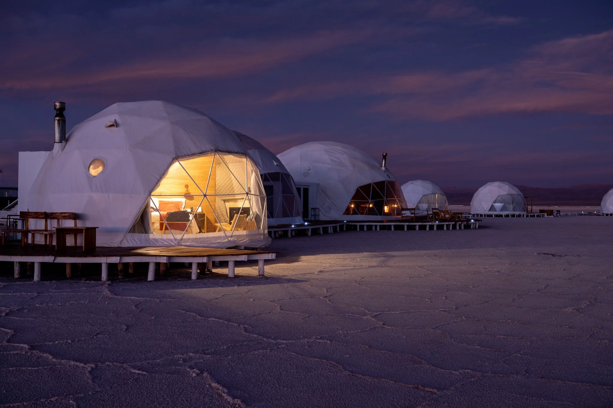 Series of geodesic domes with lit interiors on a salt flat at dusk, with a mountain range in the background.