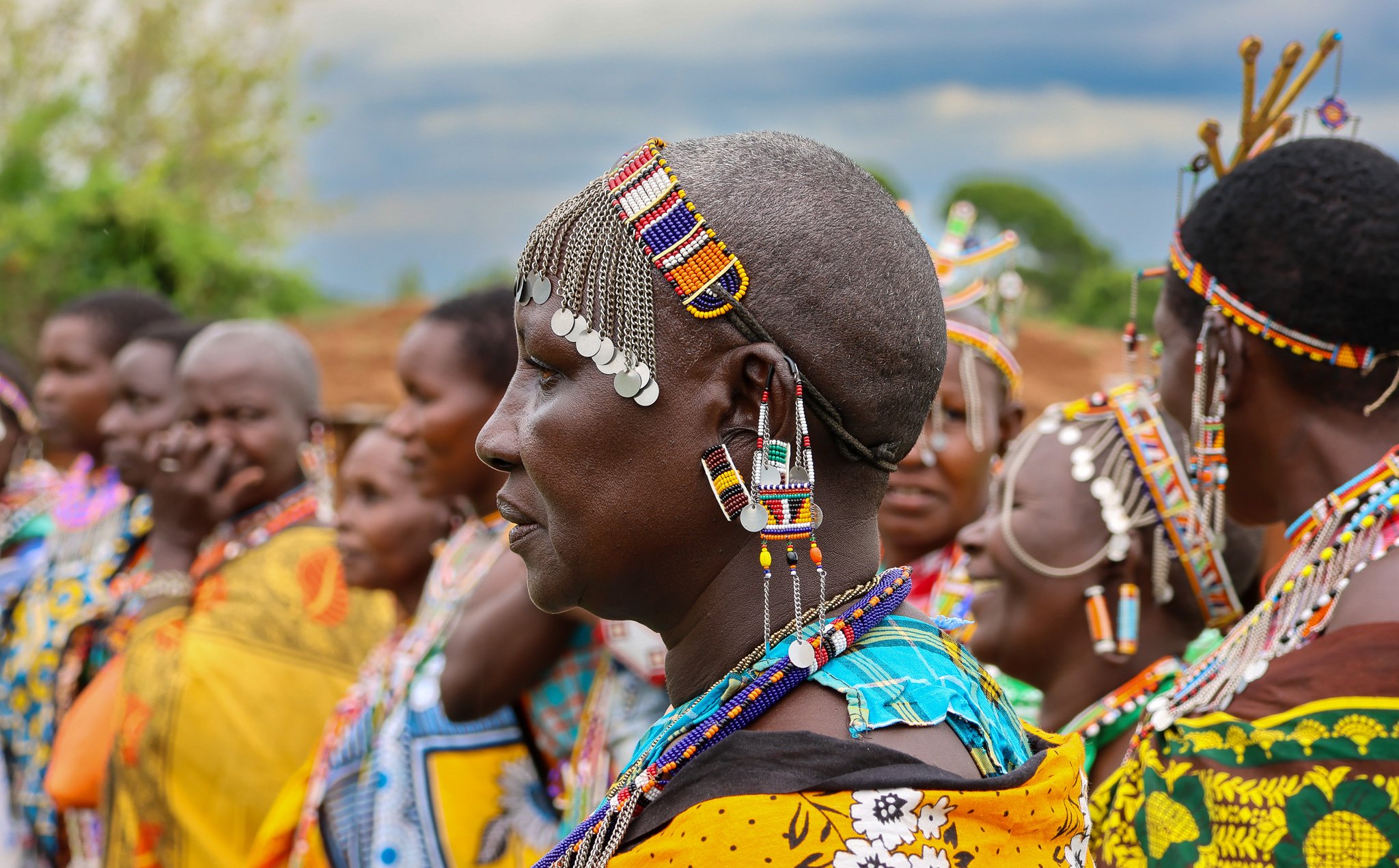 A group of Maasai women dressed in colorful traditional attire and jewelry, standing outdoors.
