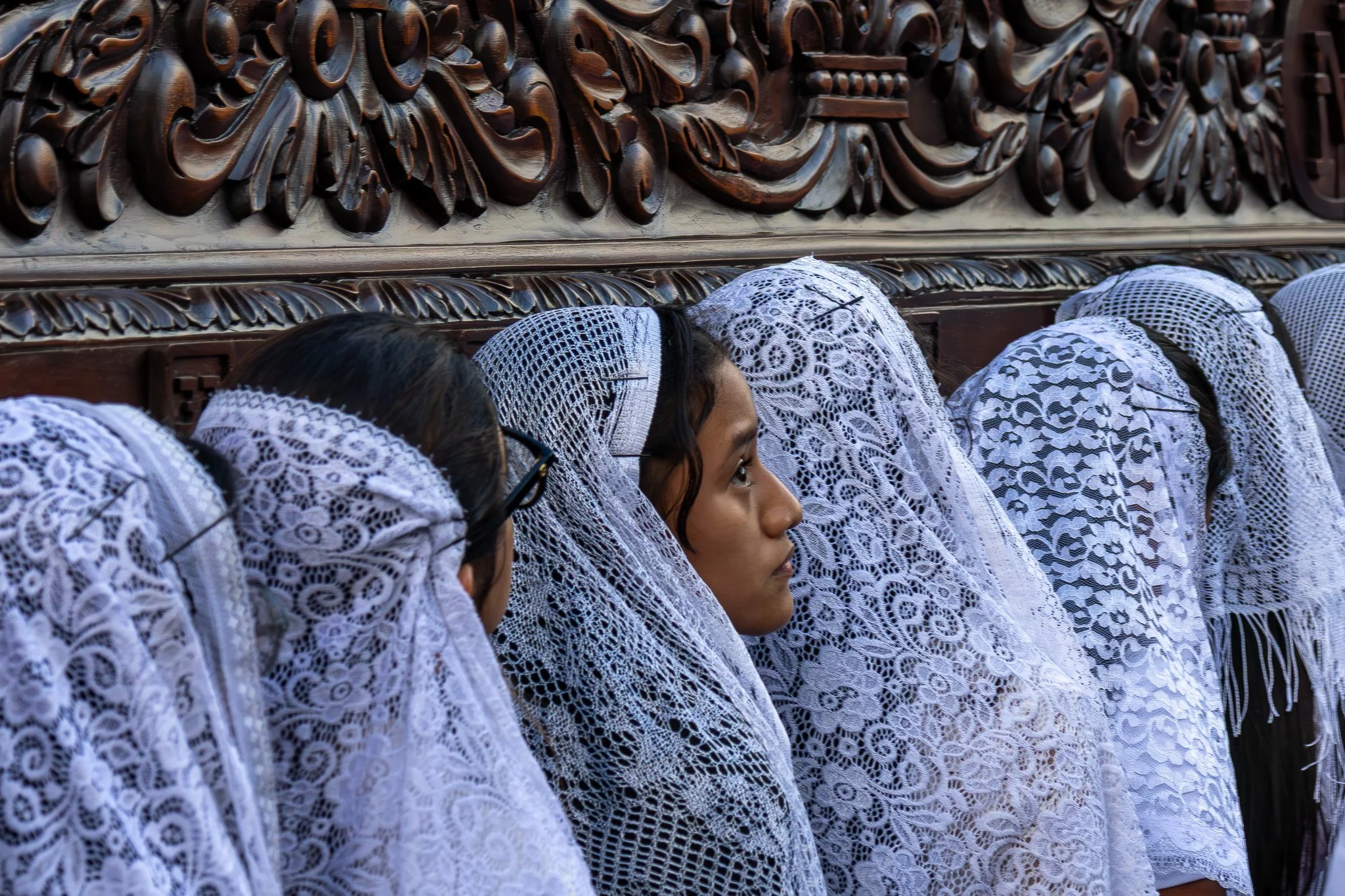A group of women with embroidered white lace head coverings sitting against a carved wooden wall.