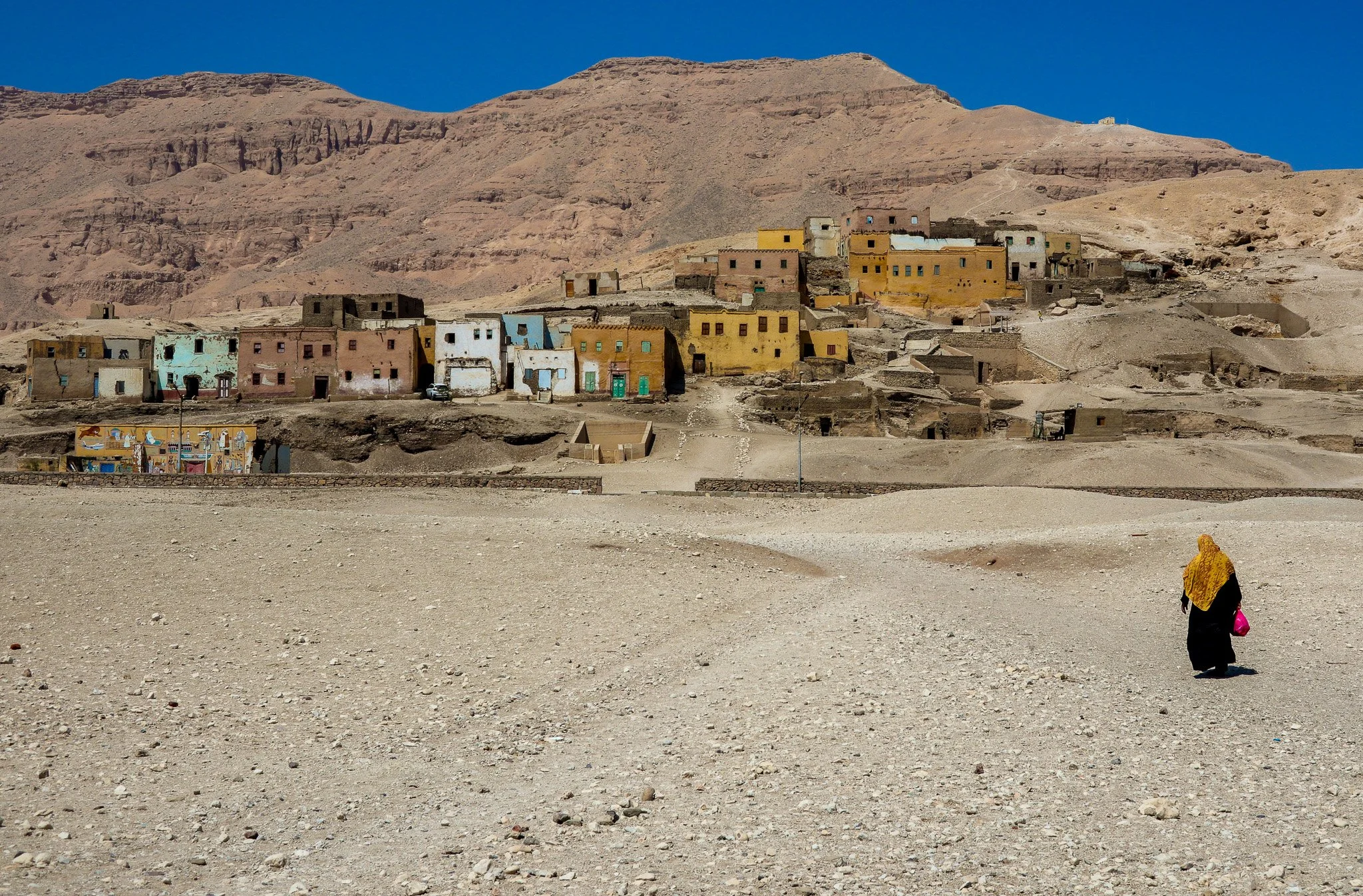 A desert landscape with a small woman wearing a yellow headscarf and a long black dress walking towards a hillside village with colorful houses under a clear blue sky.