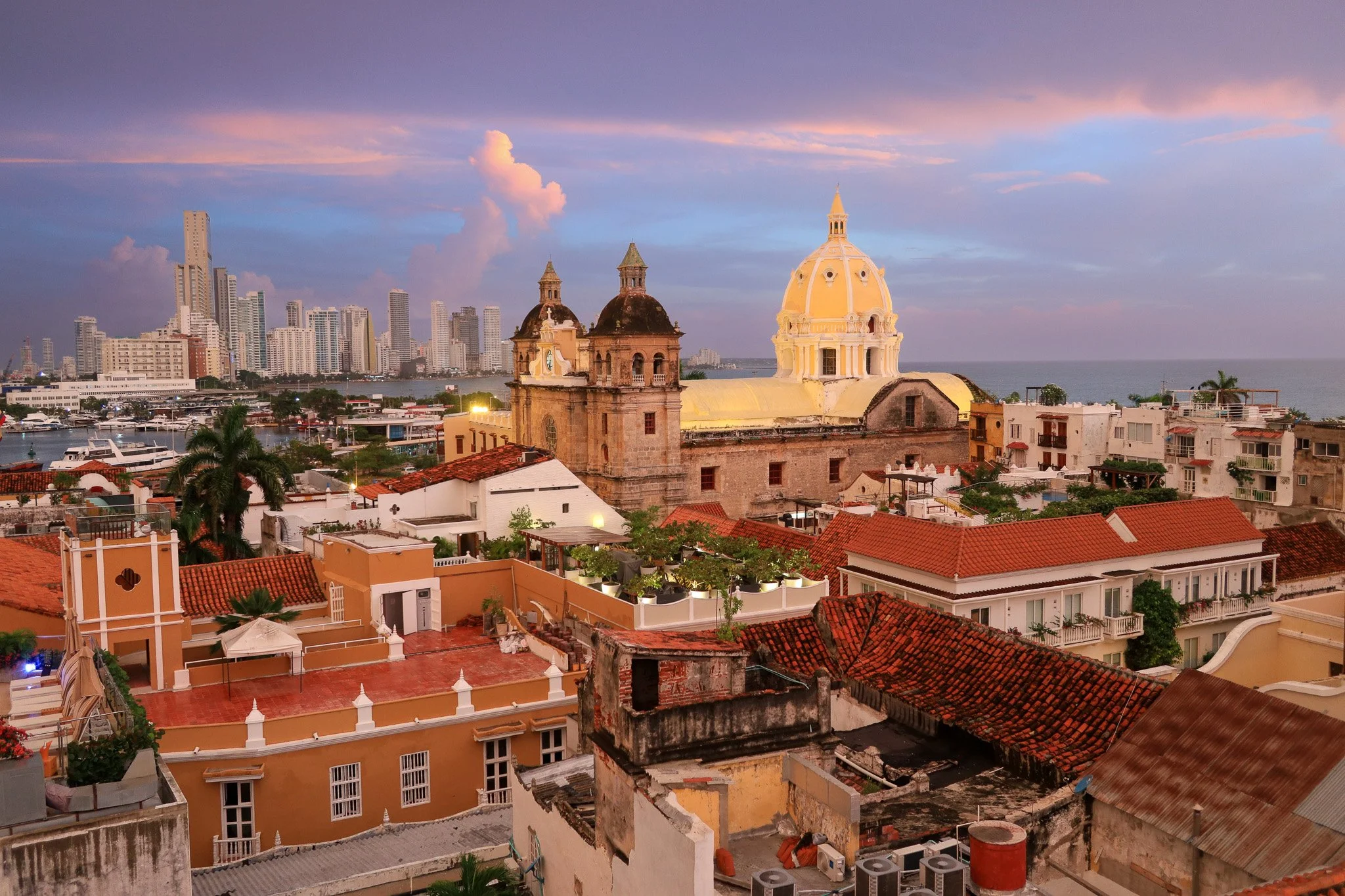 Cityscape at sunset featuring historic church with domed yellow roof and two bell towers, surrounded by colorful buildings with red tile roofs, modern high-rise buildings in the background, and a marina with yachts on the water.