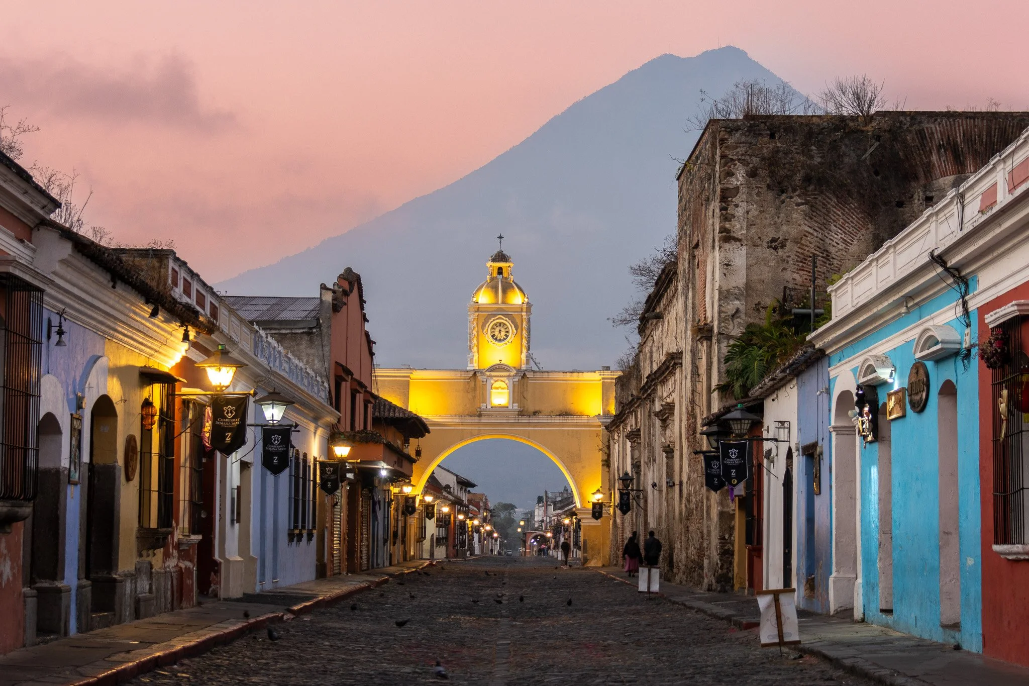 Colorful street in a historic city with a large illuminated clock tower in the background and a mountain behind it during dusk.