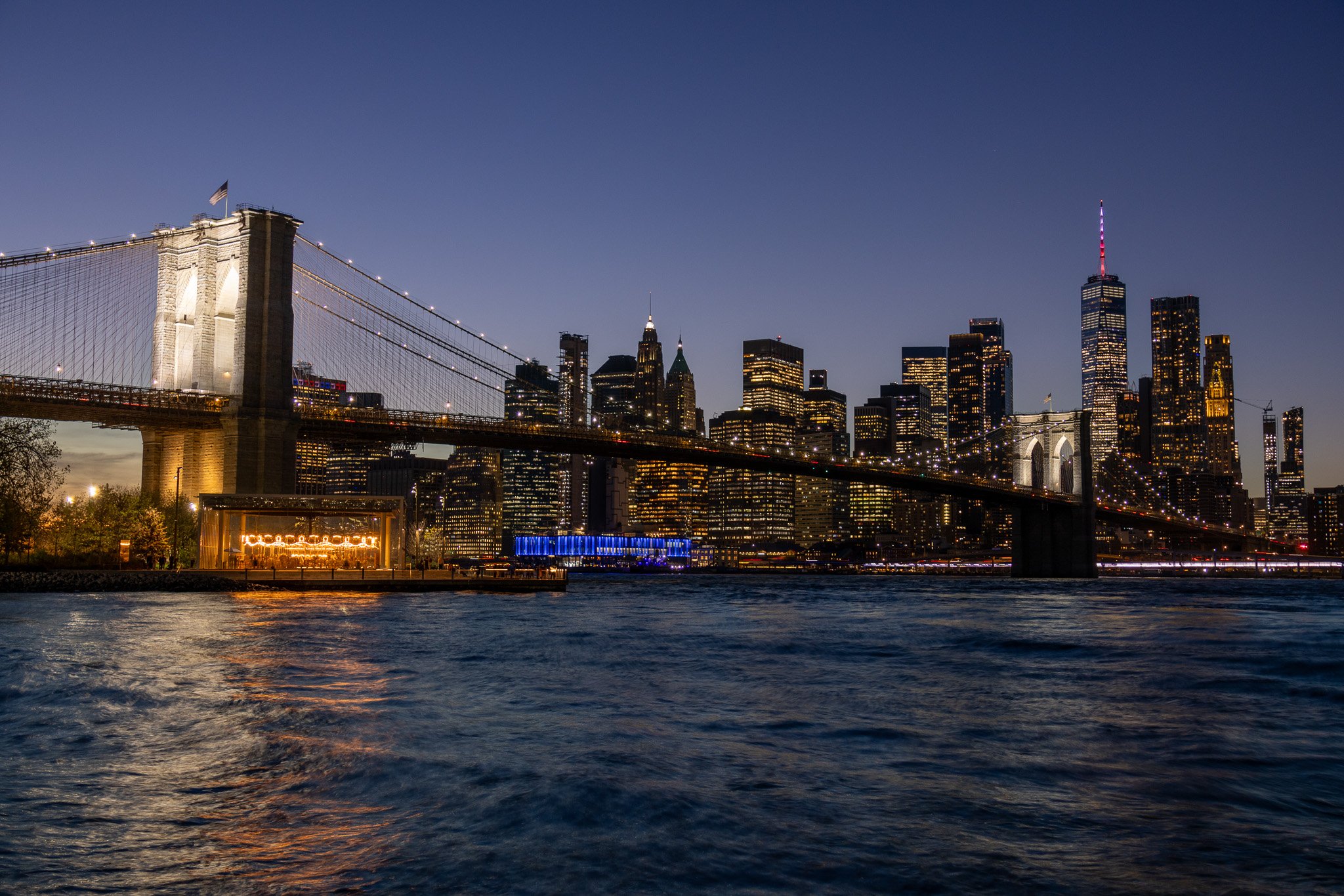 Night view of the New York City skyline with the Brooklyn Bridge in the foreground, illuminated skyscrapers, and the East River reflecting city lights.