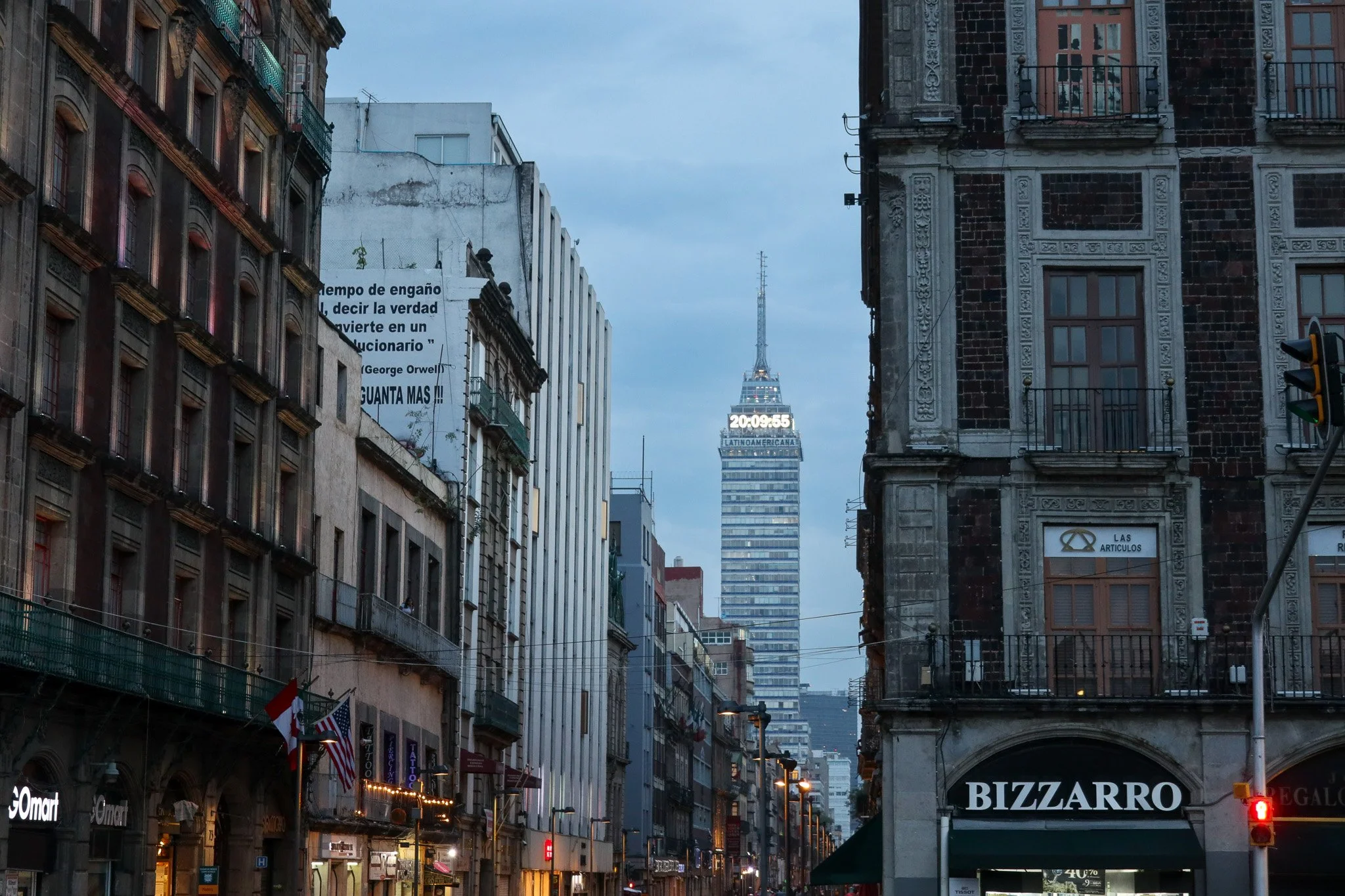 City street scene during early evening with tall buildings and the Torre Latinoamericana skyscraper in the background. Streetlights are on, and a few commercial signs and flags are visible on the buildings.