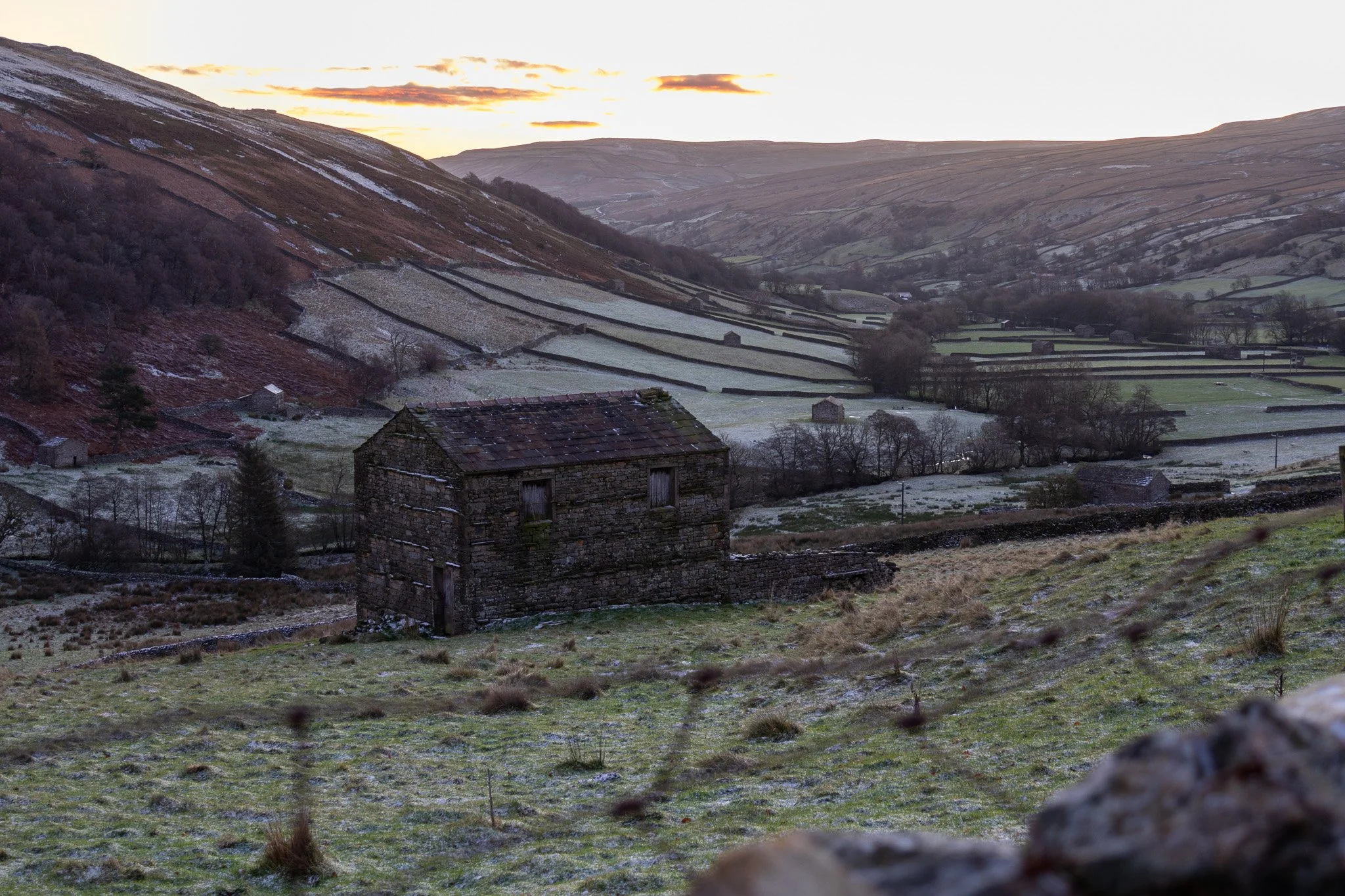 A rural landscape at sunset with rolling hills, stone fences, and an old stone barn in the foreground.
