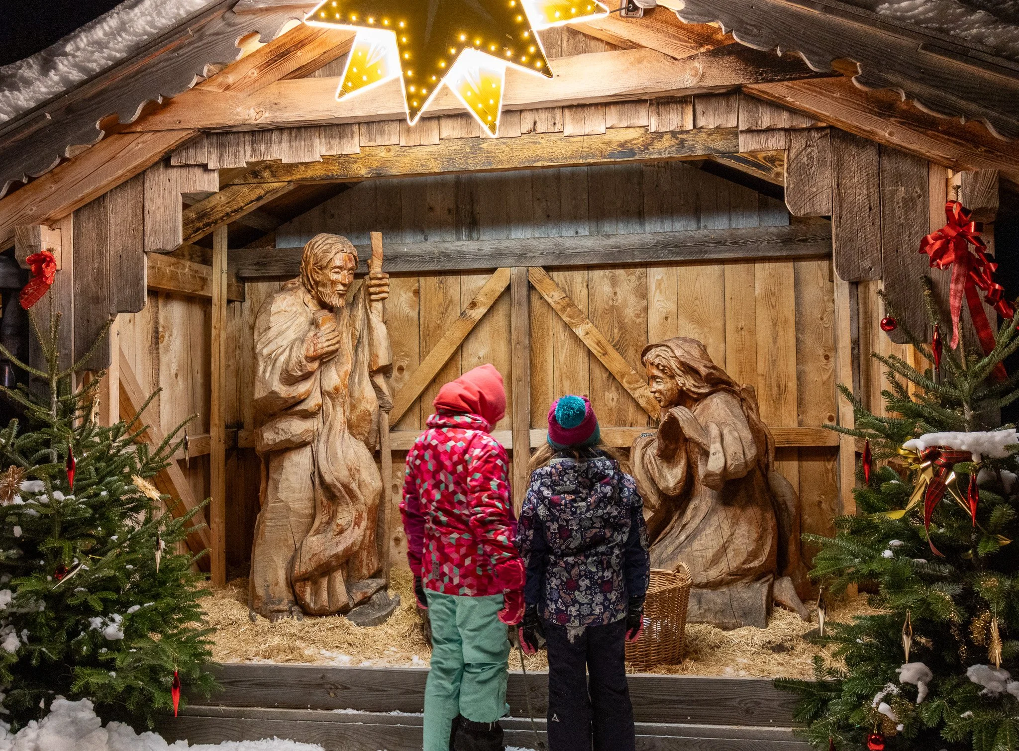 Three children dressed in colorful winter clothing and hats standing in front of a Nativity scene with wooden statues of Jesus, Mary, and Joseph inside a wooden stable with Christmas trees and decorations surrounding it.