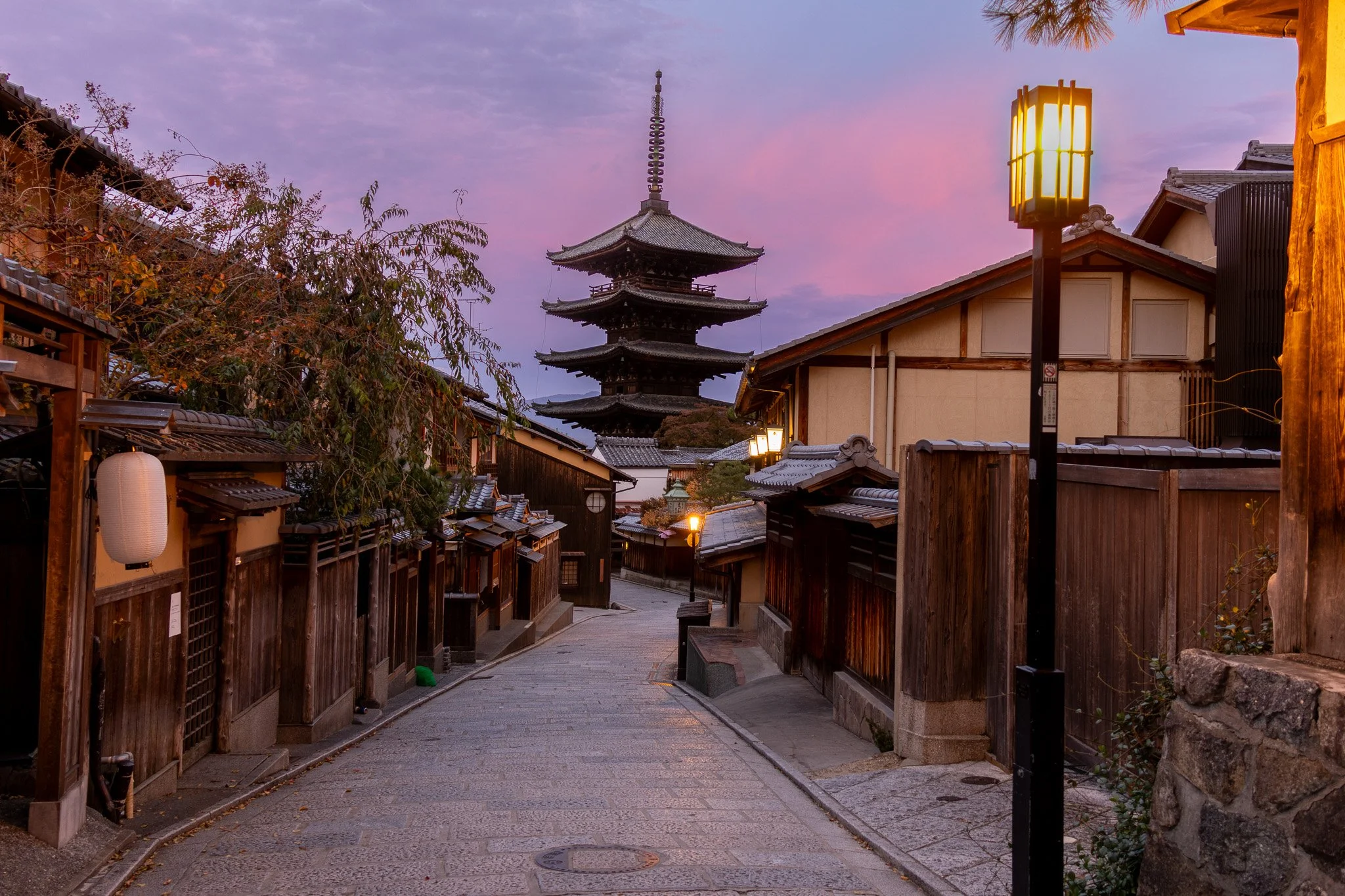 Traditional Japanese street with wooden fences and lanterns, leading to a five-story pagoda at sunset with a colorful sky.
