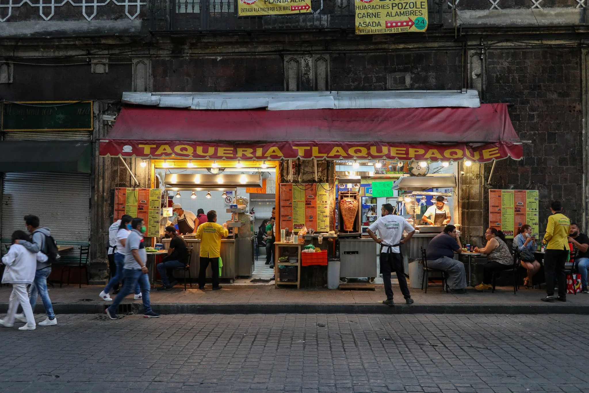 Street view of a taqueria restaurant with a red awning labeled "Taqueria Tlaquepaque." Several people, some wearing masks, are seated at tables outside, while others are standing or walking in front of the establishment. The interior is visible with 