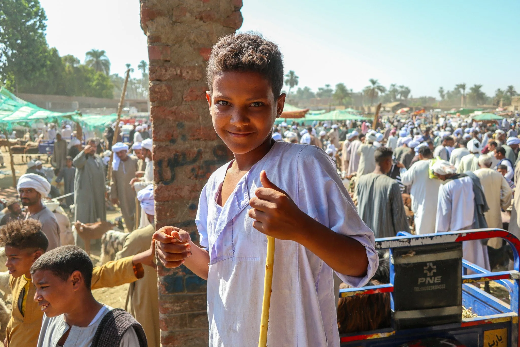 A smiling young boy in white traditional clothing holding a yellow stick, standing in front of a busy outdoor market with many people dressed in traditional attire, some wearing white head coverings, and various market stalls with green canopies in t