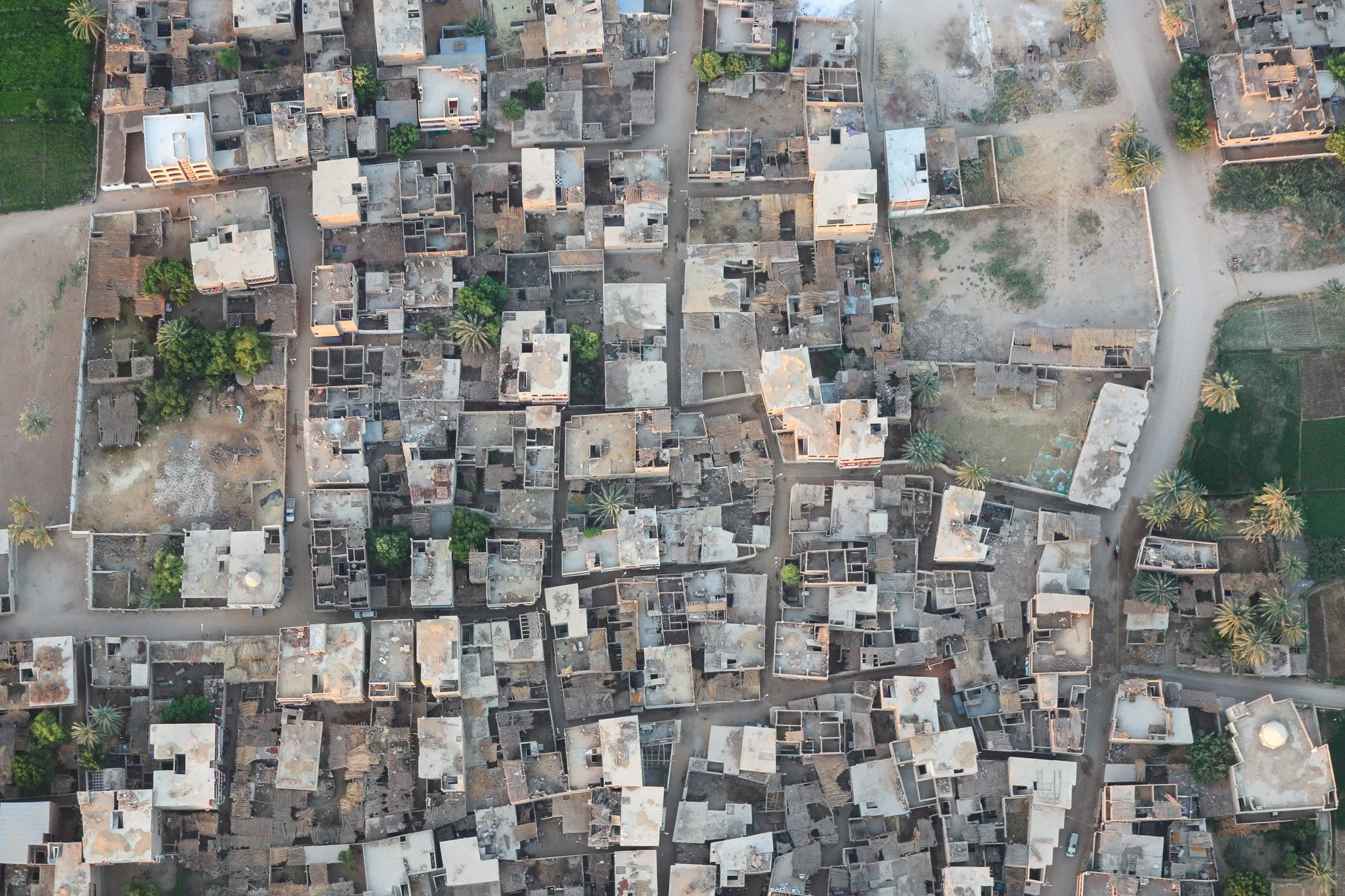An aerial view of a densely packed neighborhood showing numerous small houses with flat roofs, some with trees and open spaces around them, and dirt roads between the buildings.