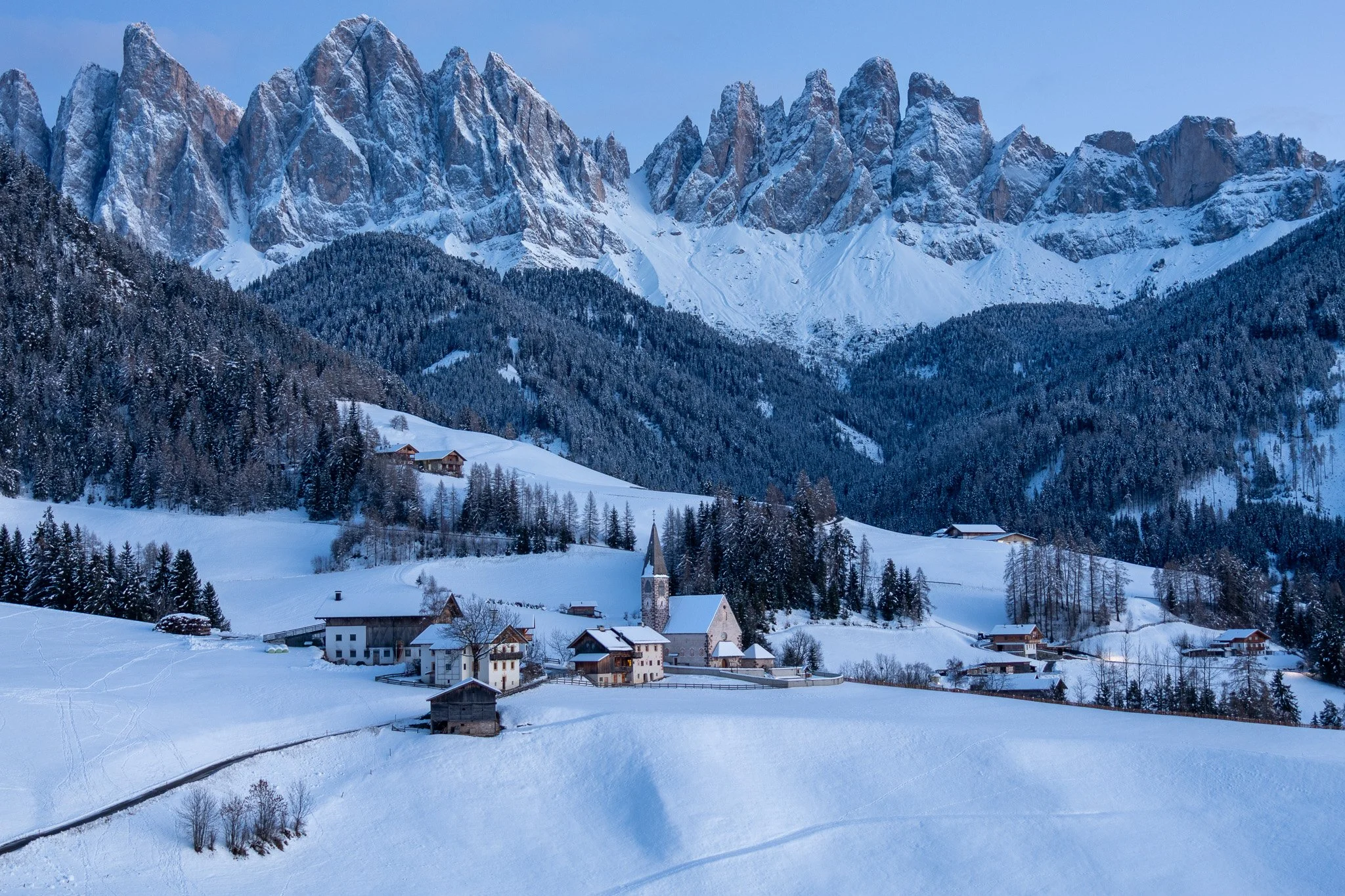 Snow-covered village in a valley with mountains in the background, featuring a church with a tall steeple and scattered houses surrounded by trees.