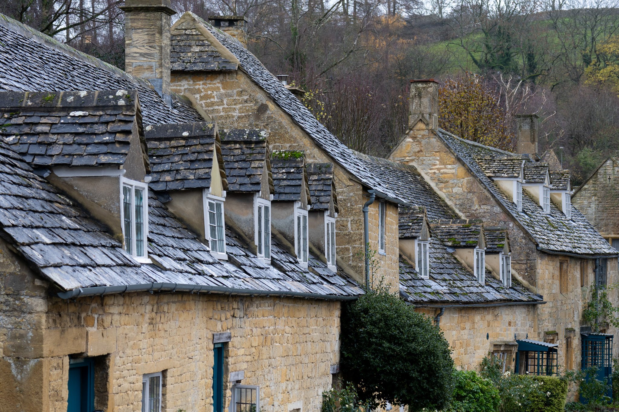 Row of old stone houses with slate roofs, chimneys, and small dormer windows, surrounded by trees and greenery in a rural setting.
