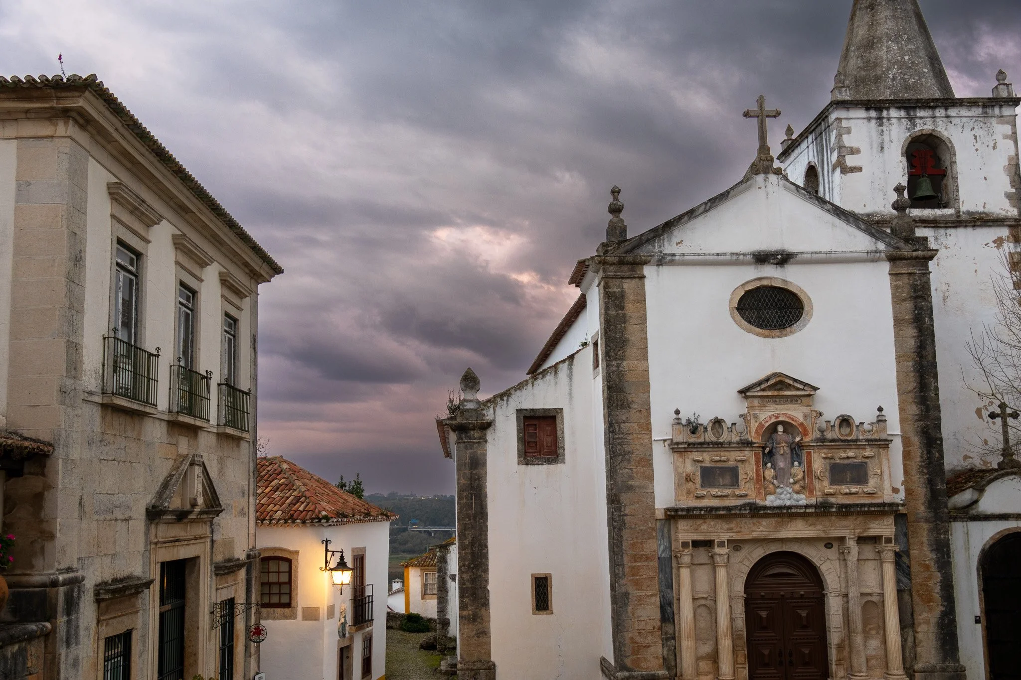 Old stone and white church buildings with a cloudy sky and a streetlamp at sunset.