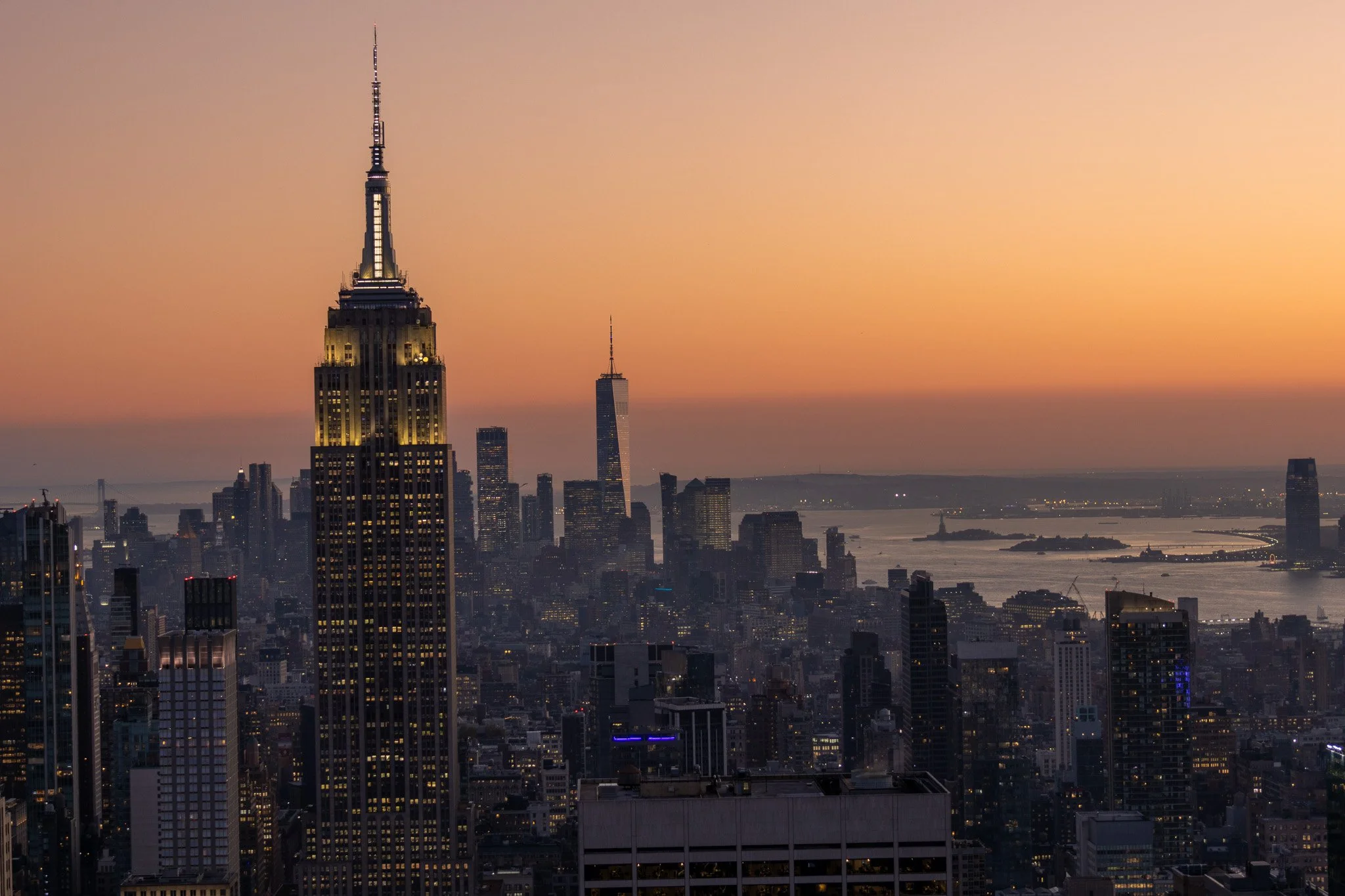 City skyline at sunset with illuminated Empire State Building in the foreground and One World Trade Center in the background, overlooking the Hudson River.