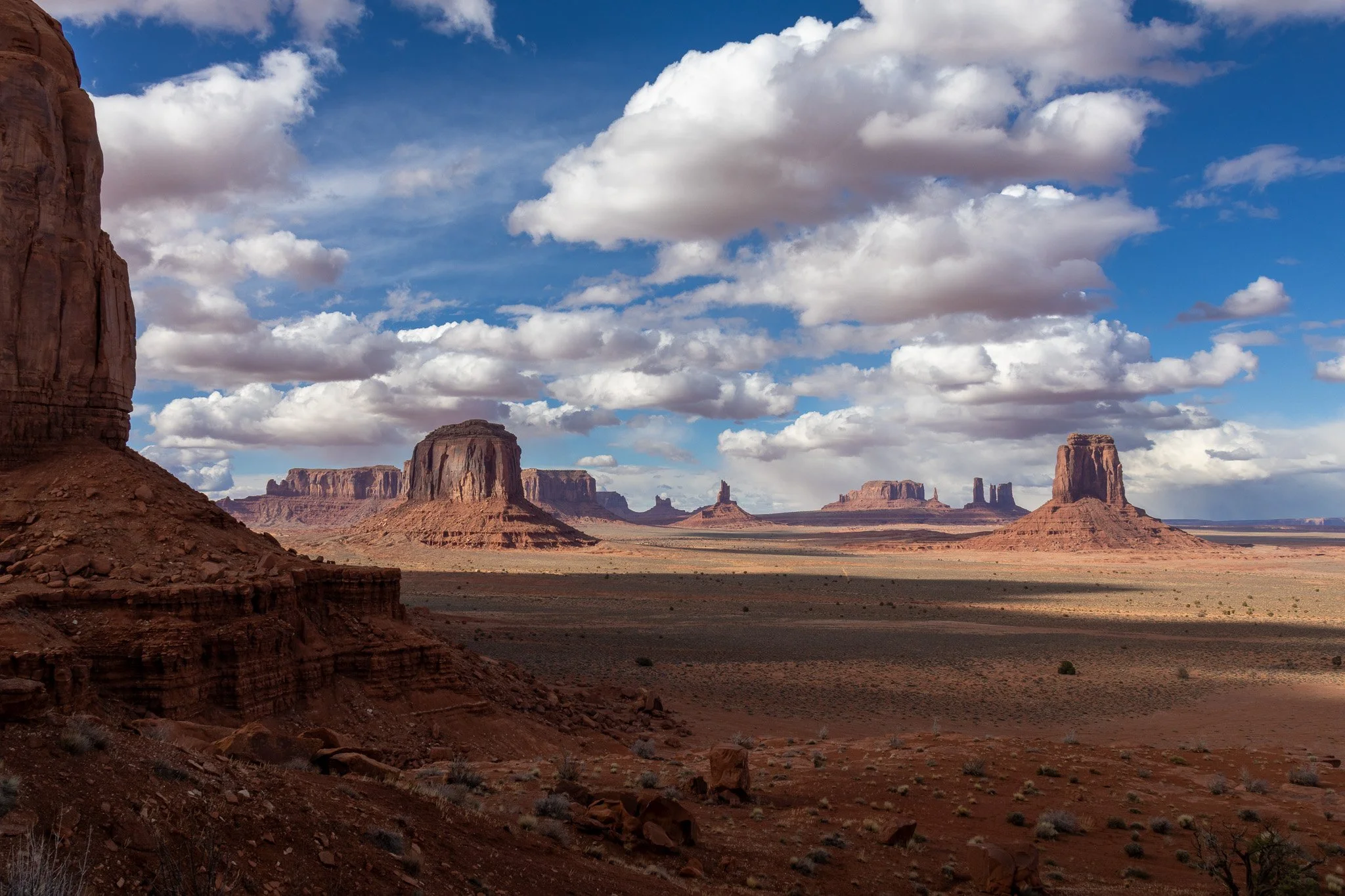 A desert landscape with large red rock formations and buttes, under a partly cloudy sky.