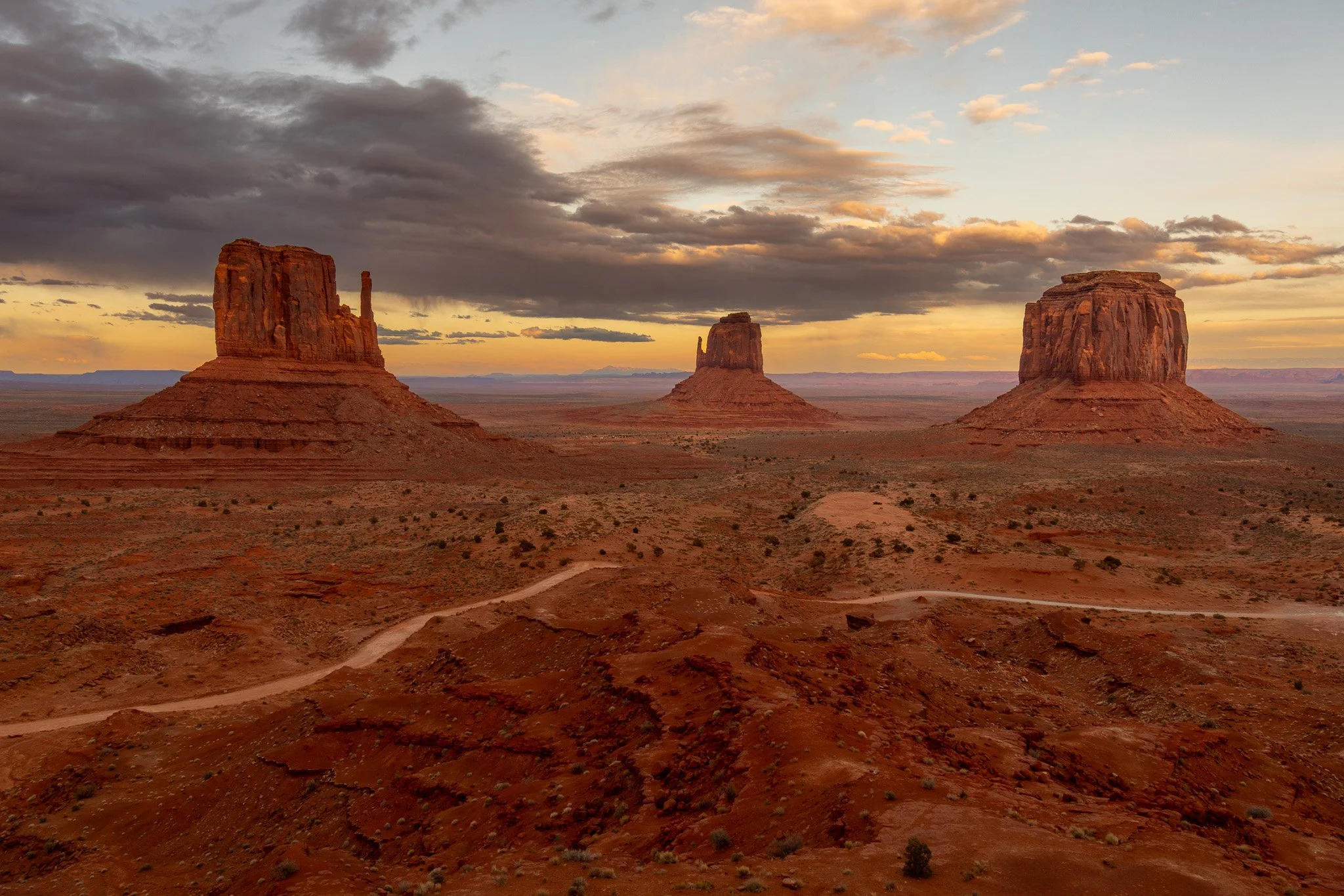 Sunset over Monument Valley with three large, flat-topped sandstone buttes rising from the desert landscape, under a sky with dark clouds and soft orange light.