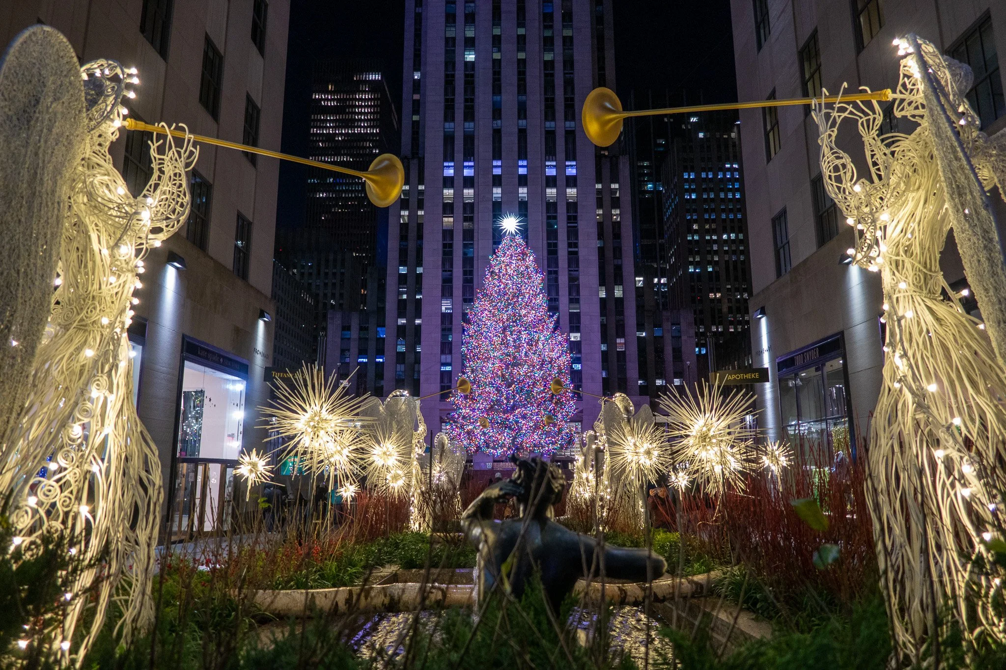 A Christmas tree decorated with multicolored lights and topped with a star, surrounded by glowing angel and star-shaped lights, in a city plaza at night.