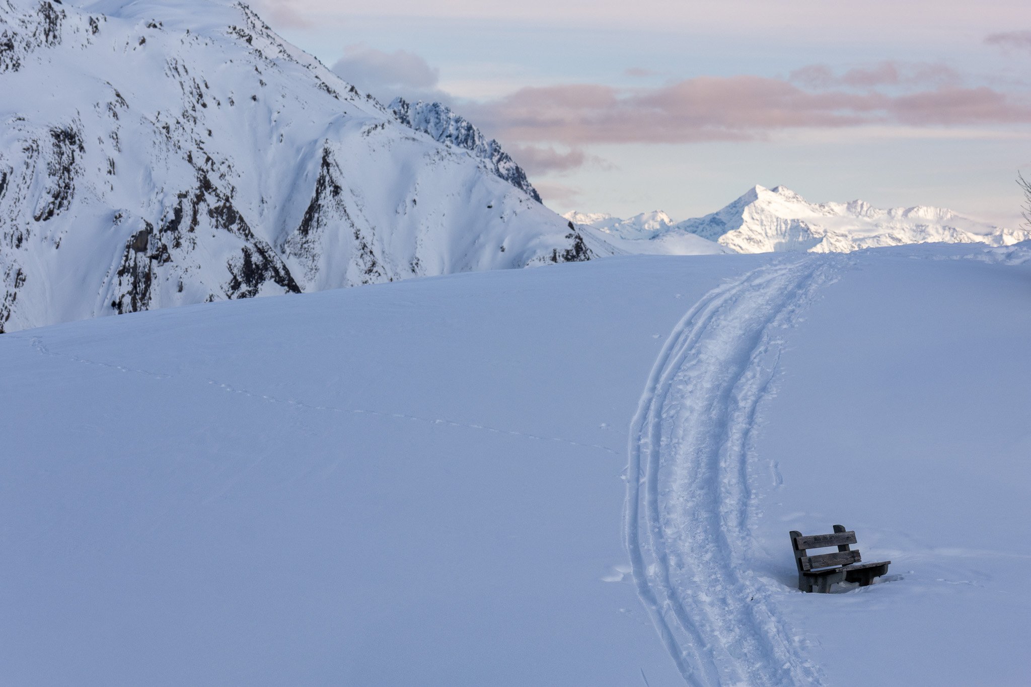Snow-covered mountain landscape with ski tracks and a bench, distant mountains under a partly cloudy sky.