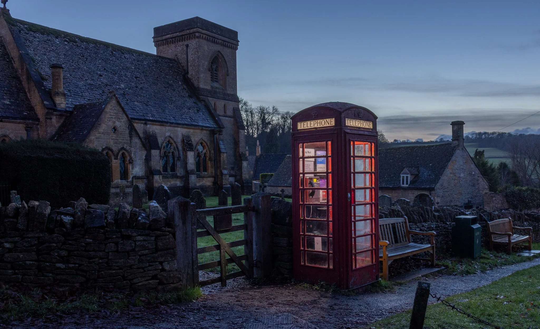 A red British telephone booth near a stone church and benches in a rural village during dusk.