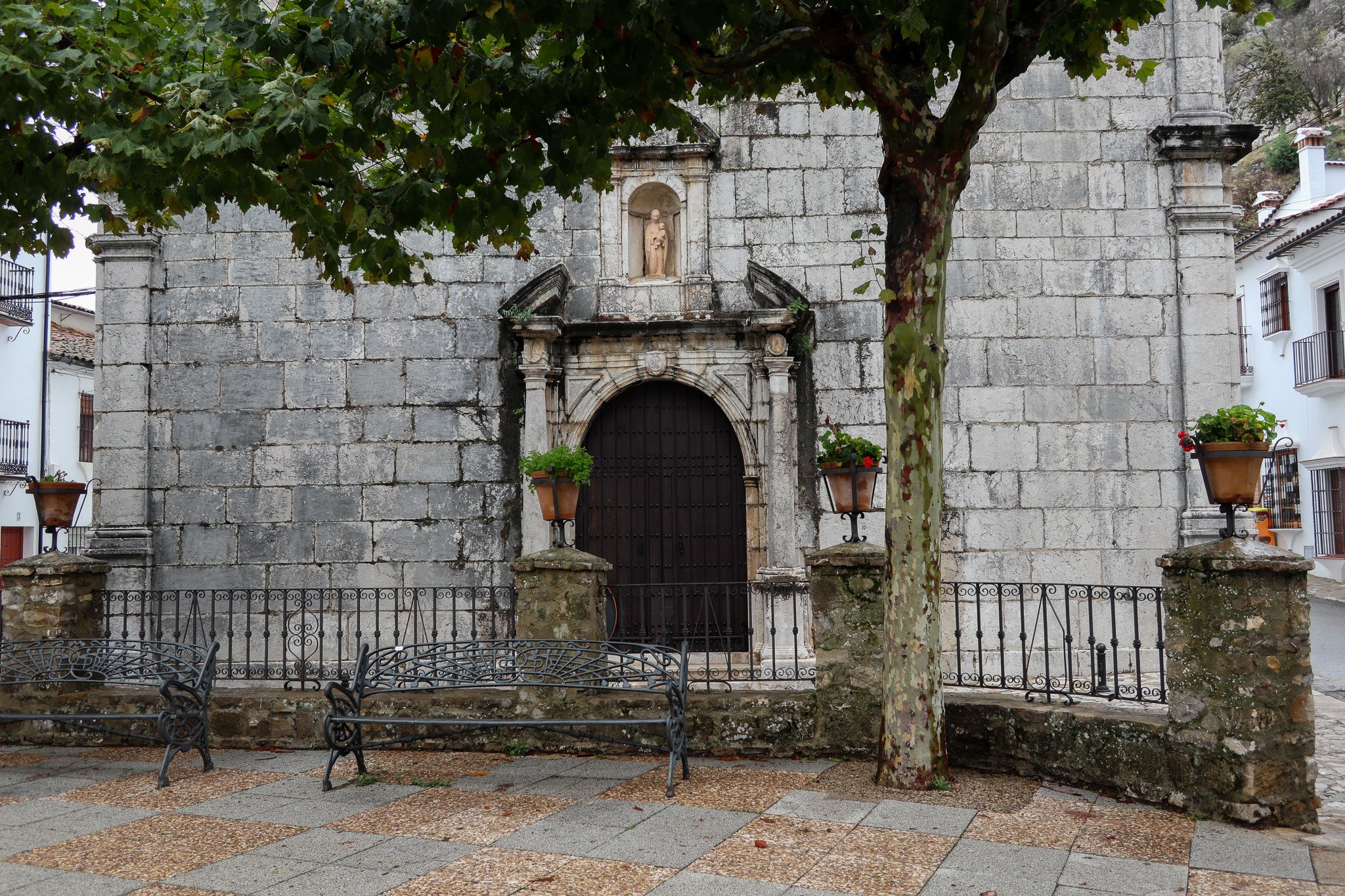 A historic stone building with an arched wooden door and decorative stonework, surrounded by a small courtyard with benches, potted plants, and a large tree with green leaves.