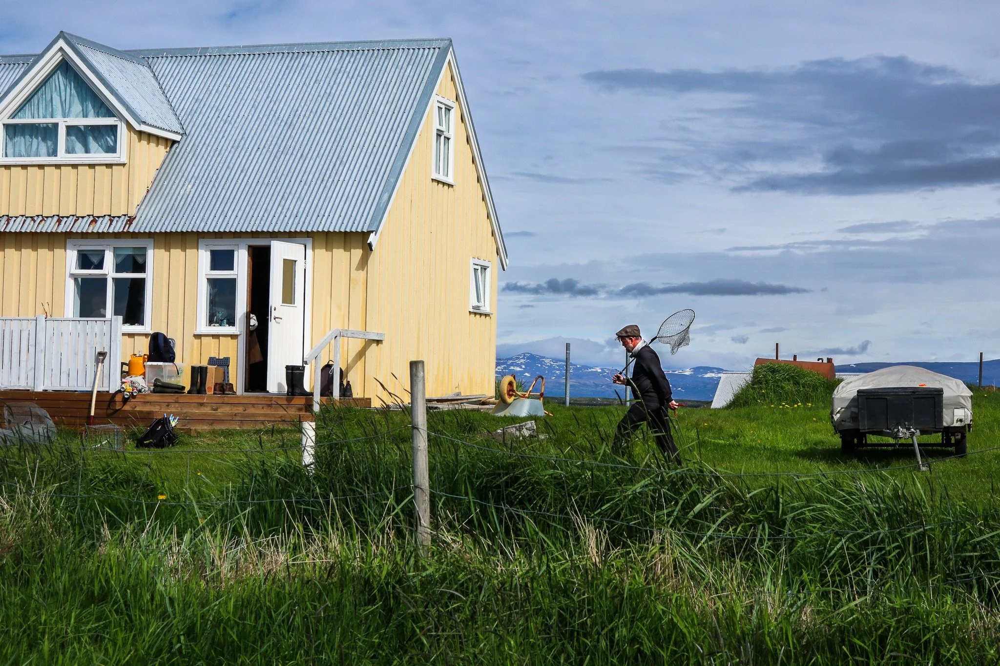 A yellow house with a blue metal roof on green grass. A man in a suit with a butterfly net walks across the yard. There are clouds in the sky and mountains in the background.