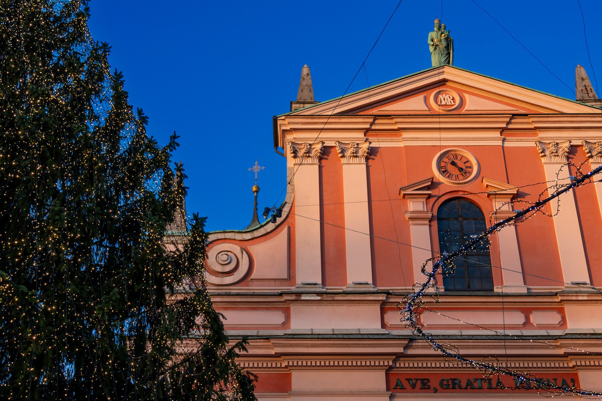 A Christmas tree decorated with lights next to a pink baroque-style church with a clock, a statue on the roof, and string lights, during evening.
