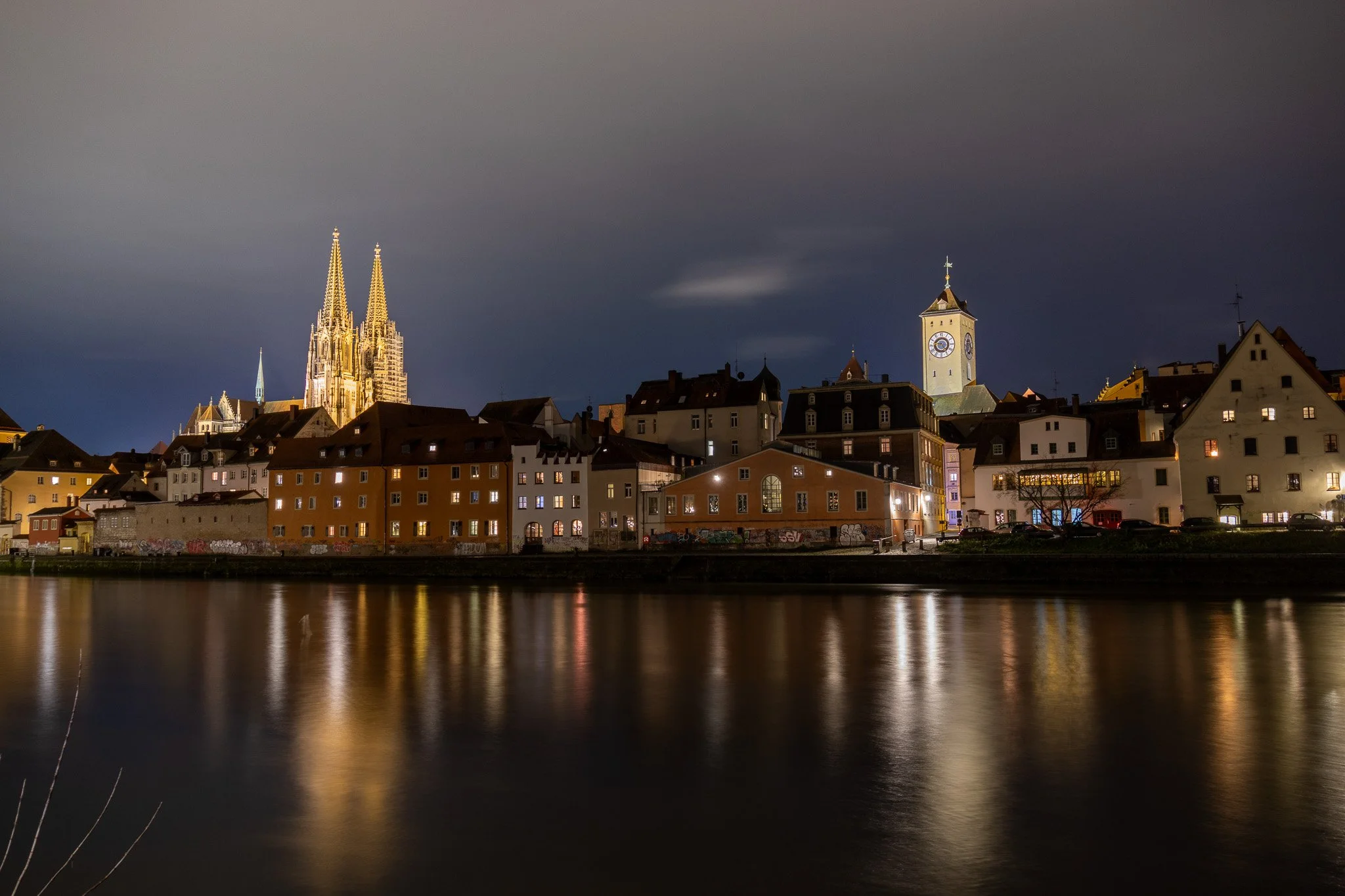 Nighttime view of a European city with a river in the foreground, historic buildings along the riverbank, and a large cathedral with tall spires illuminated in the background.