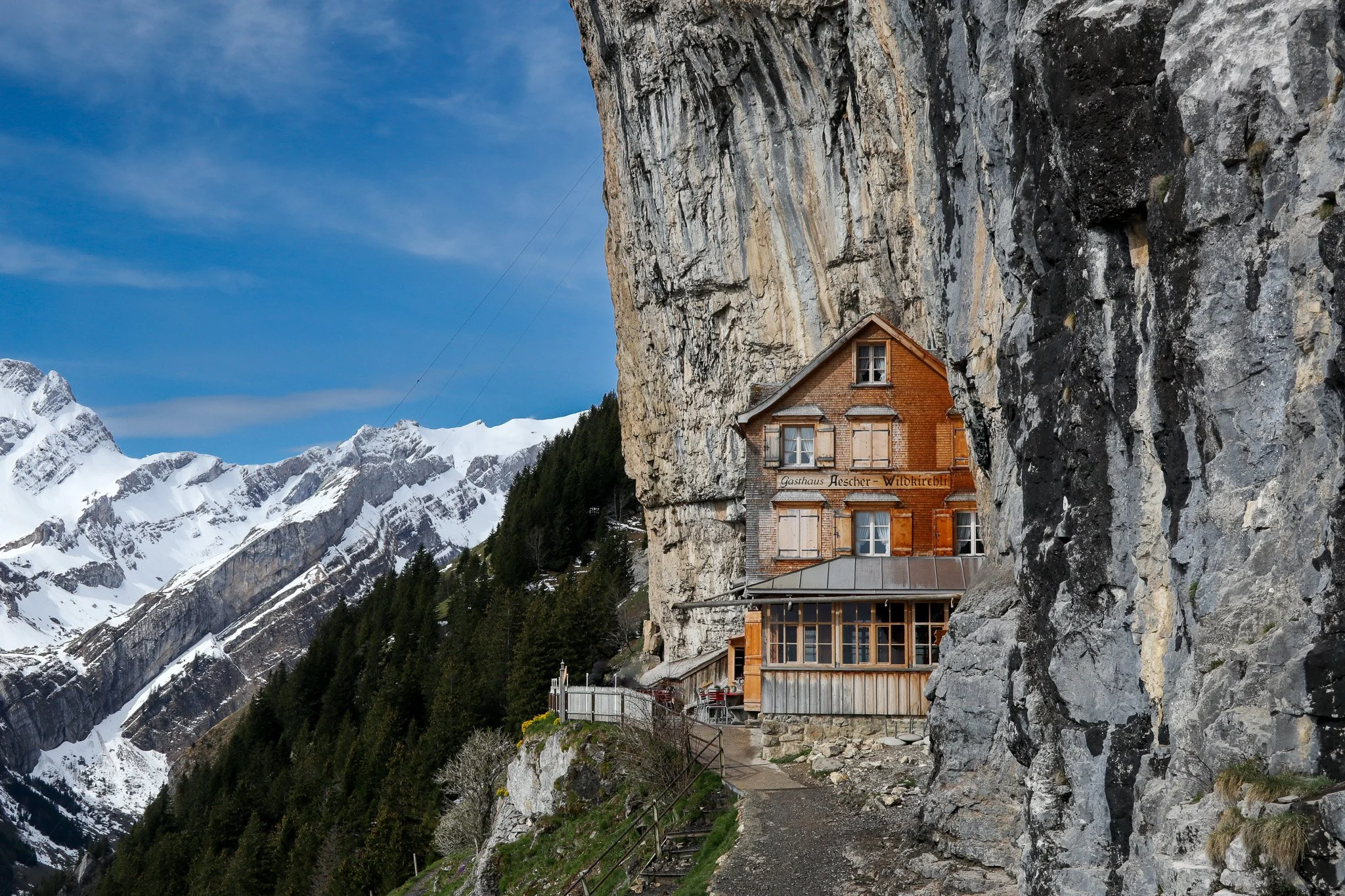 A rustic wooden house built into a rock face on a mountainside, with snow-capped peaks in the background and a cloudy blue sky above.