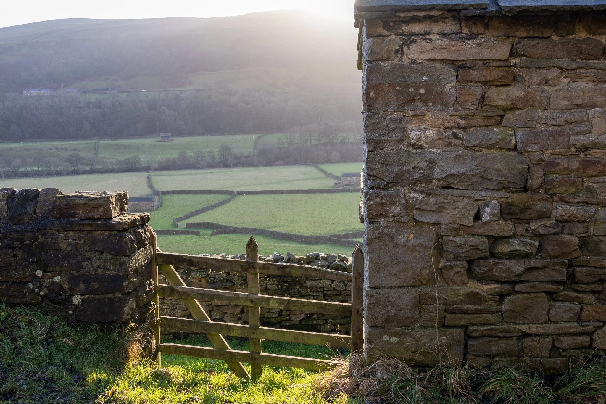 View of a countryside with green fields, stone fences, and distant hills, seen through an opening between a stone wall and an old wooden gate at sunset.