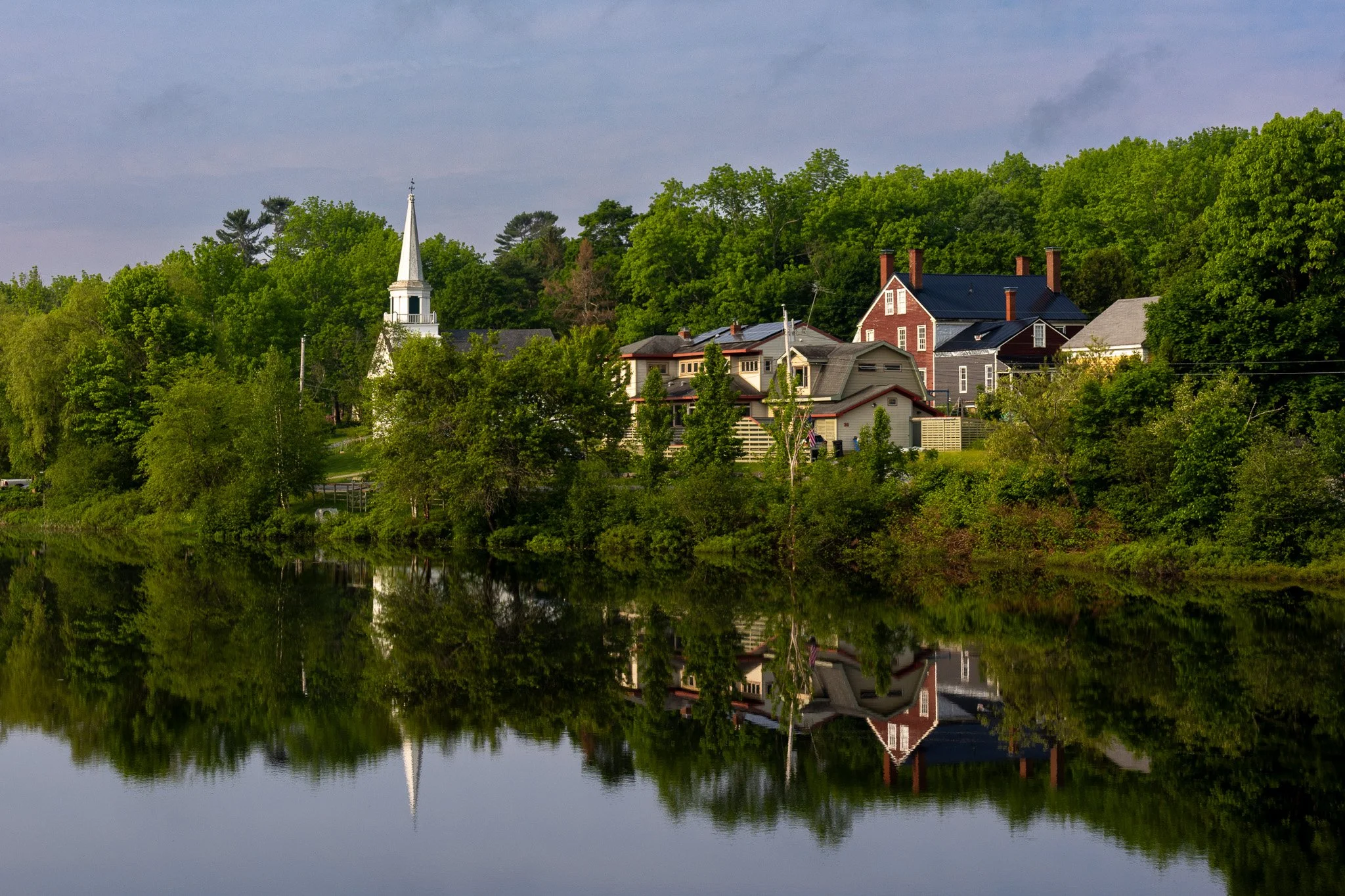 A scenic view of houses, a church with a steeple, and lush green trees along a river, with their reflection visible in the water.