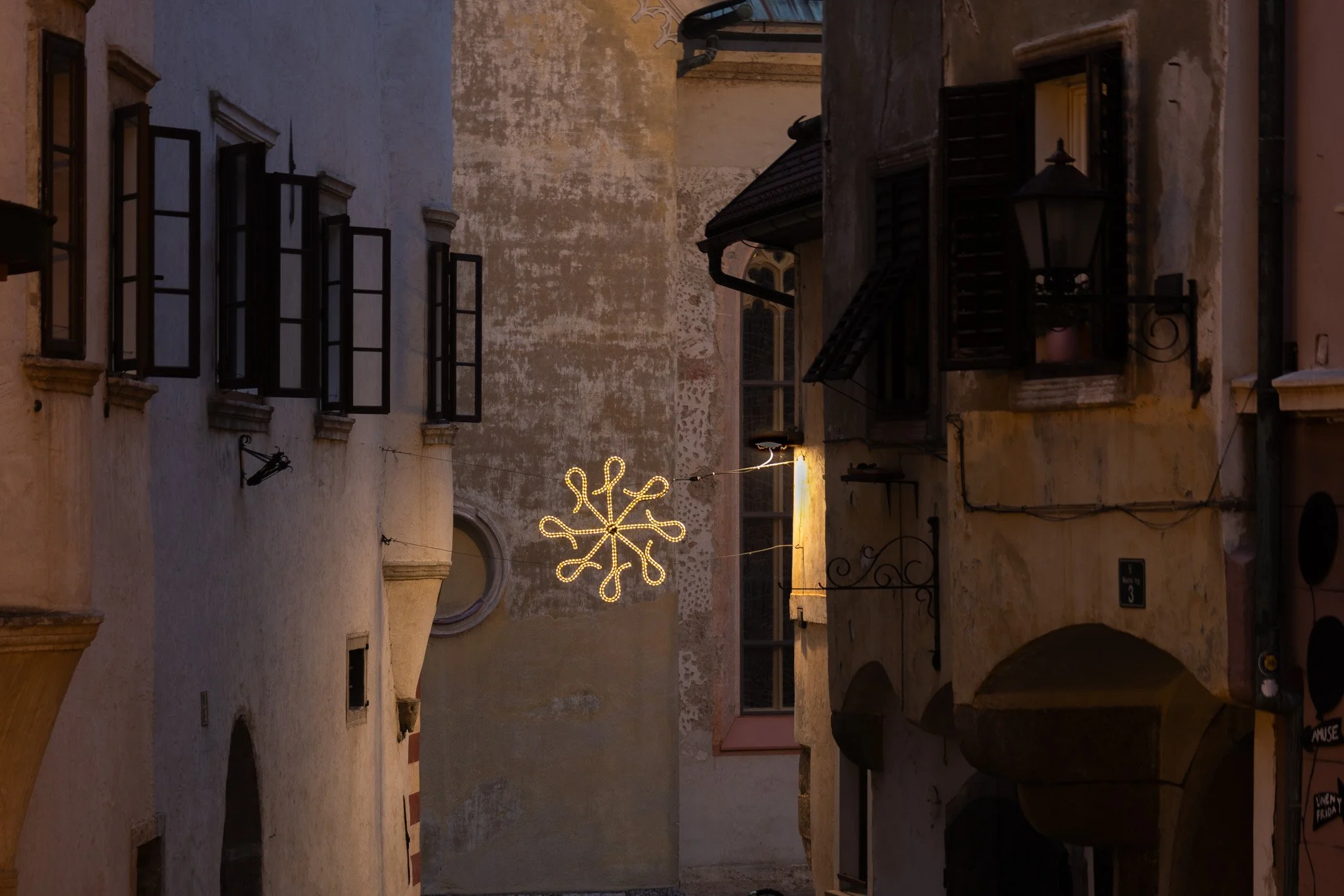 Narrow street with old buildings and a decorative holiday light in the shape of a snowflake hanging between the buildings.