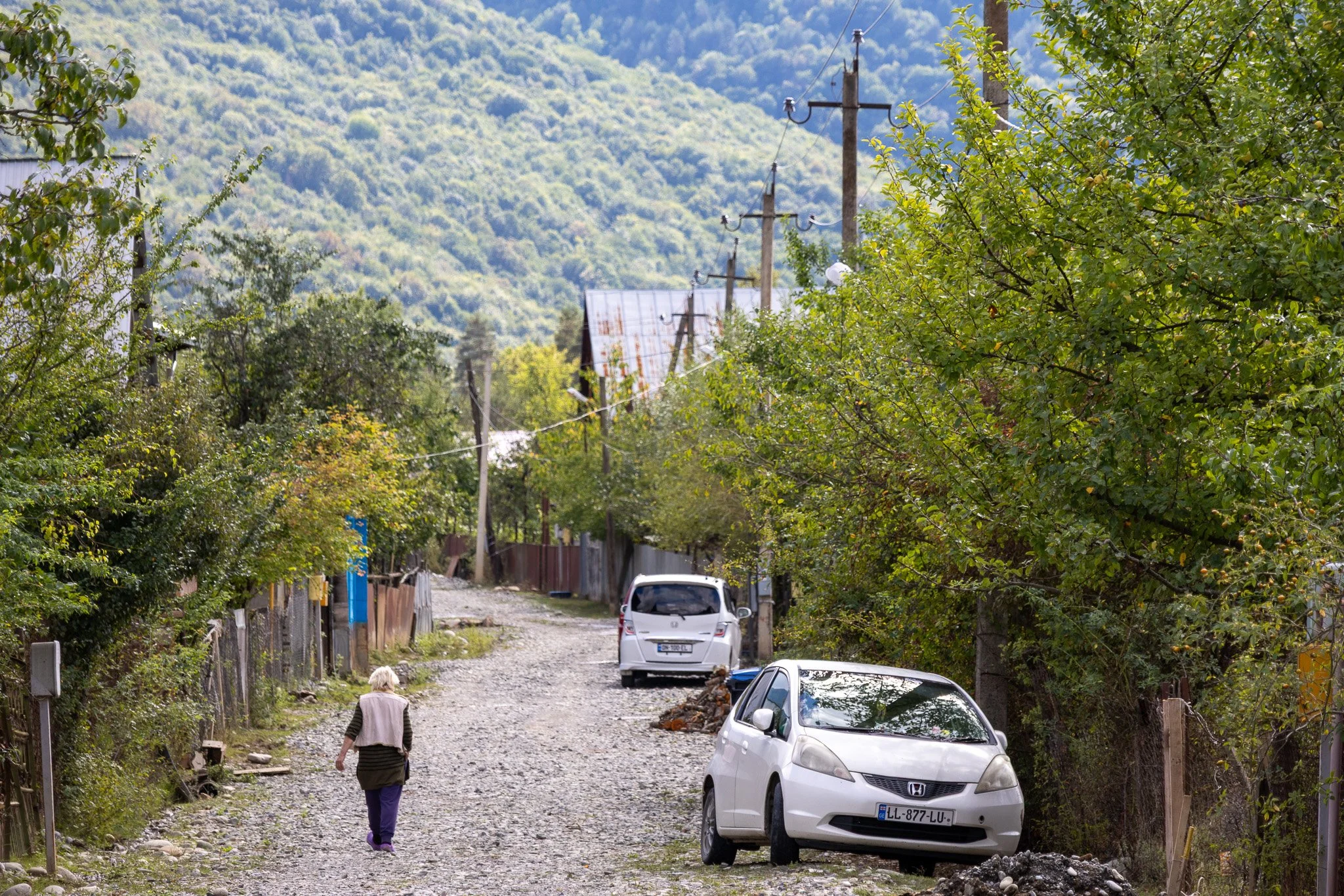 A woman walks along a gravel street in a rural area with trees and old houses. There are parked cars, including a white Honda, and power poles with wires, with a green, hilly background.