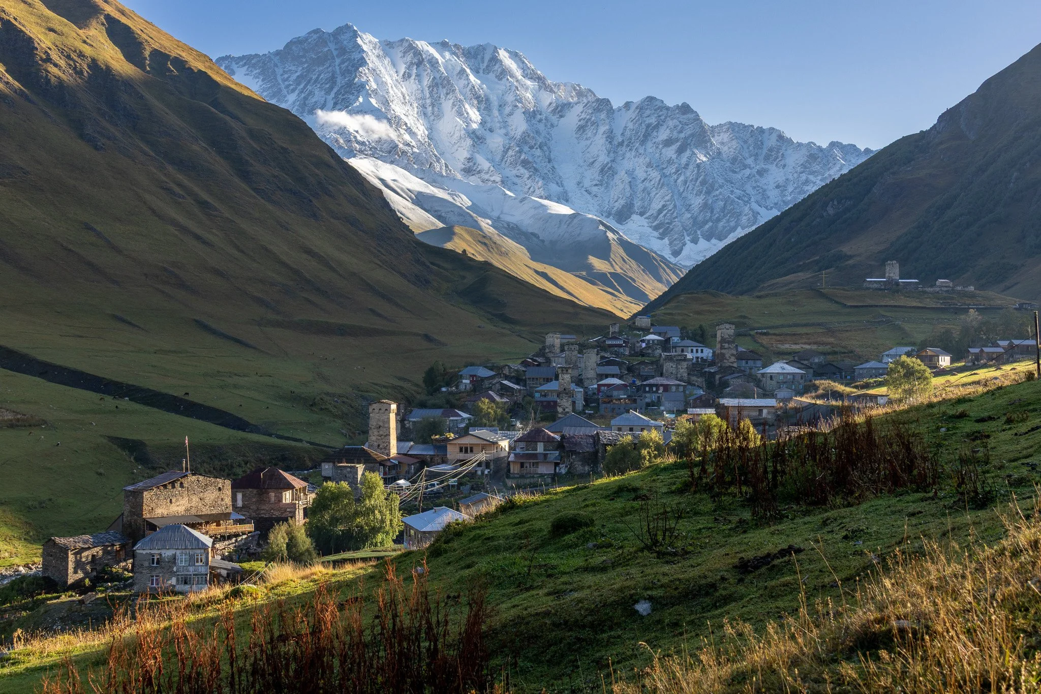 A scenic mountain village nestled in green hills with snow-capped mountains in the background.