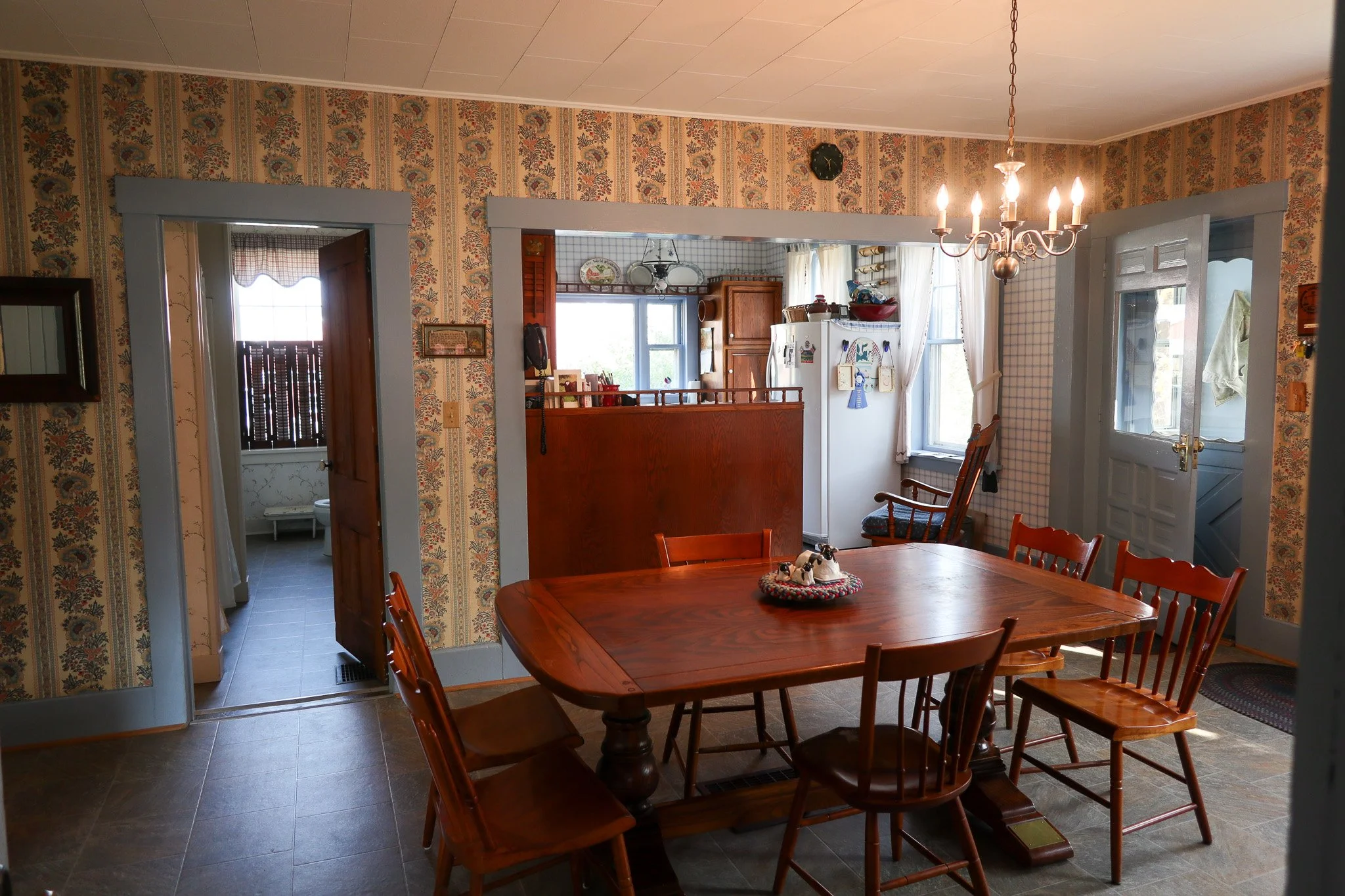 A dining room with wood table and chairs, wallpapered walls, a chandelier, and a view into a kitchen with a refrigerator and window.