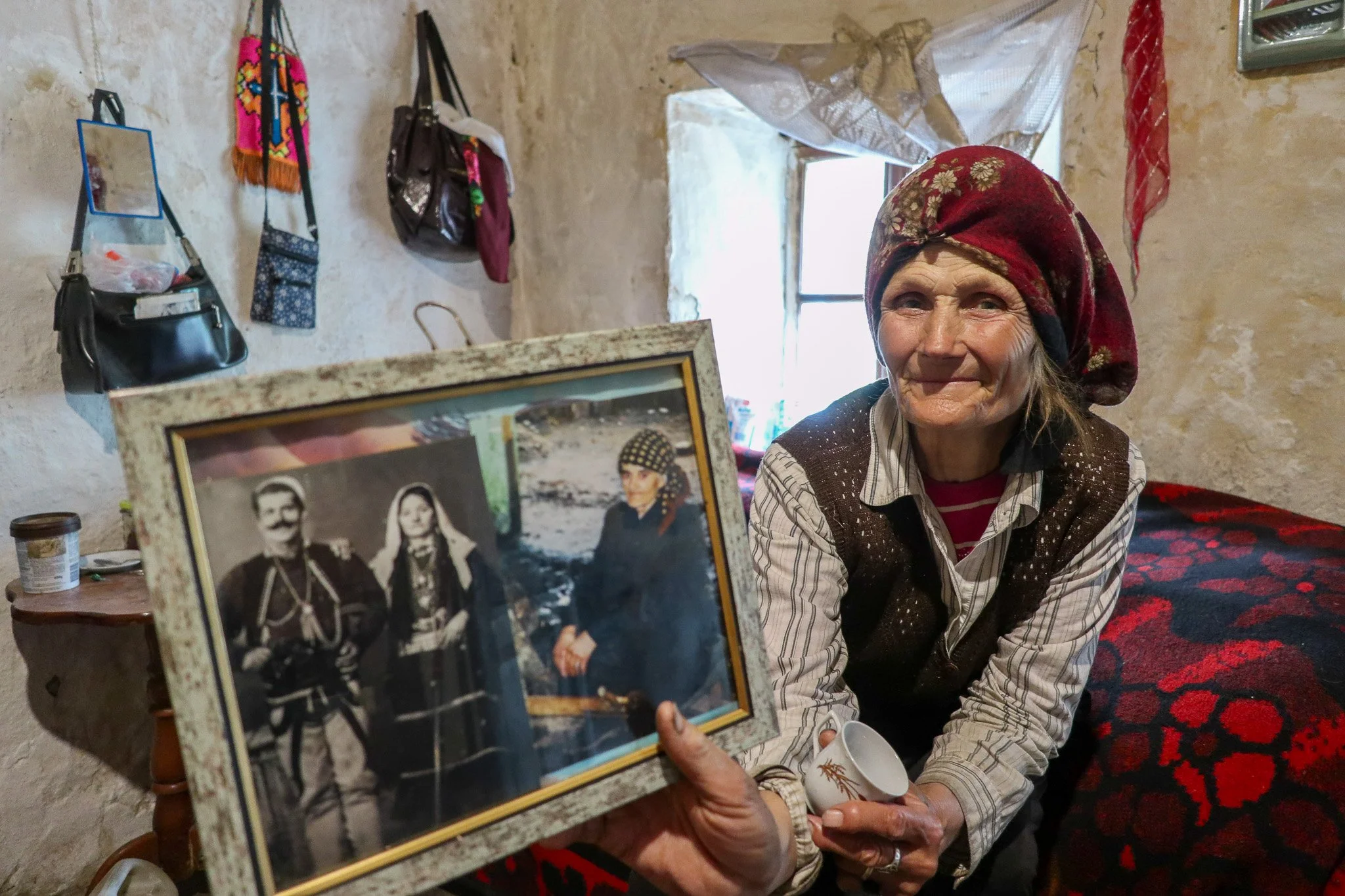 An elderly woman with a red headscarf, smiling, sitting indoors beside a wall with bags hanging on it, holding a framed black-and-white photo of two men and a woman, with a colorful blanket in the background.