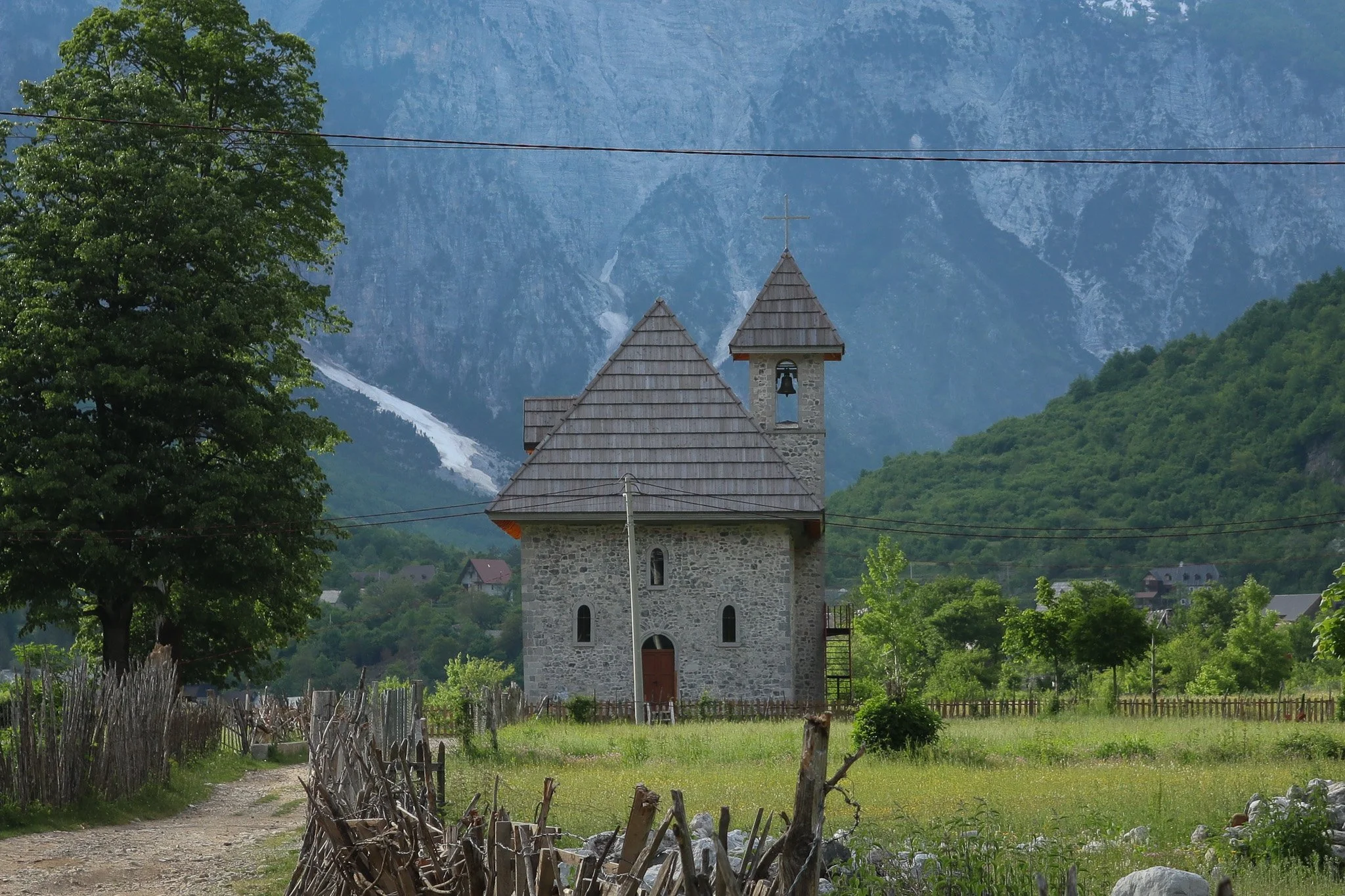 A stone church with a wooden roof and bell tower set in a green field, with trees, mountains, and a blue sky in the background.