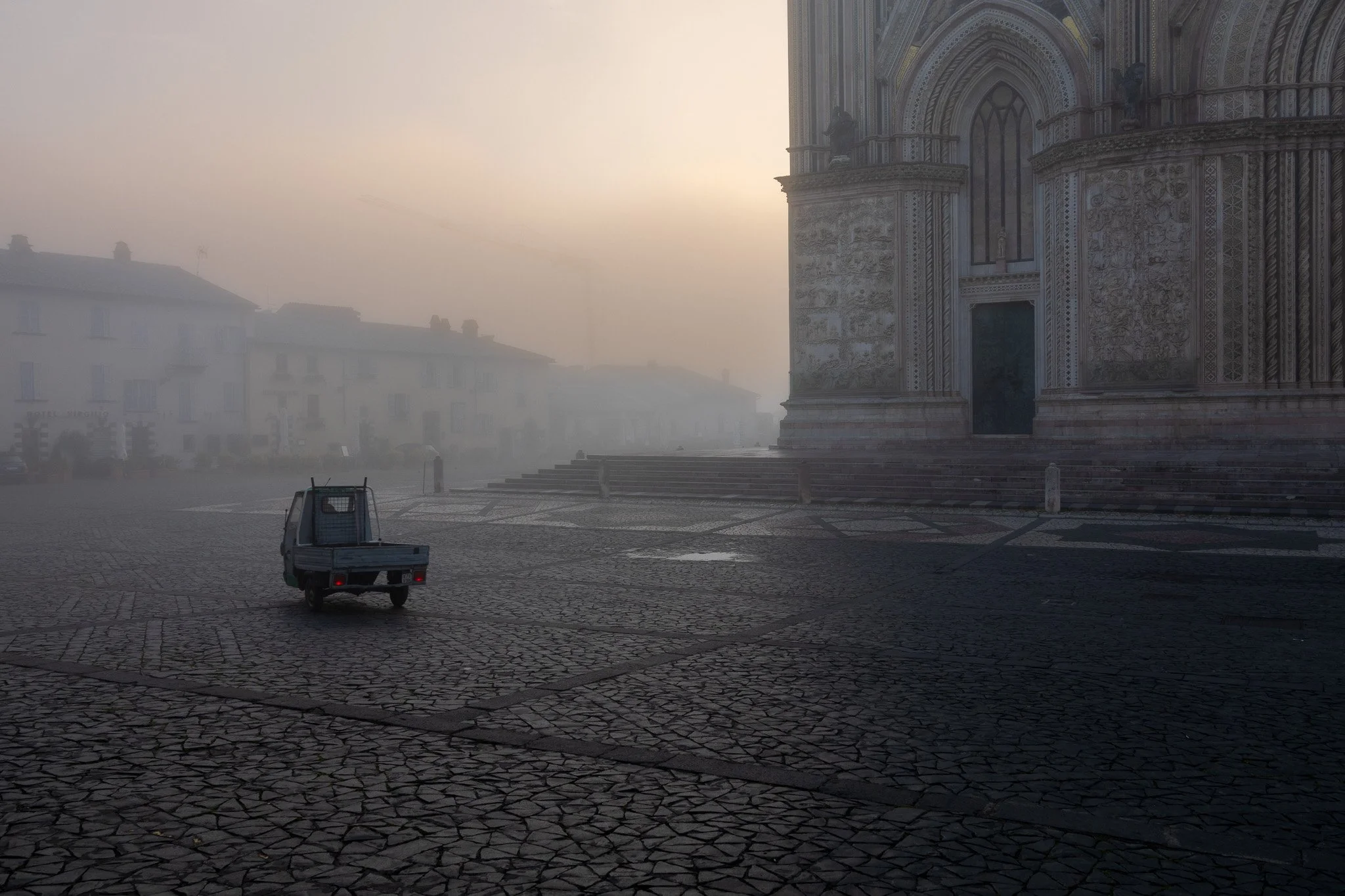Foggy square with cobblestone paving, a small vehicle, and a detailed historic building facade in the background.