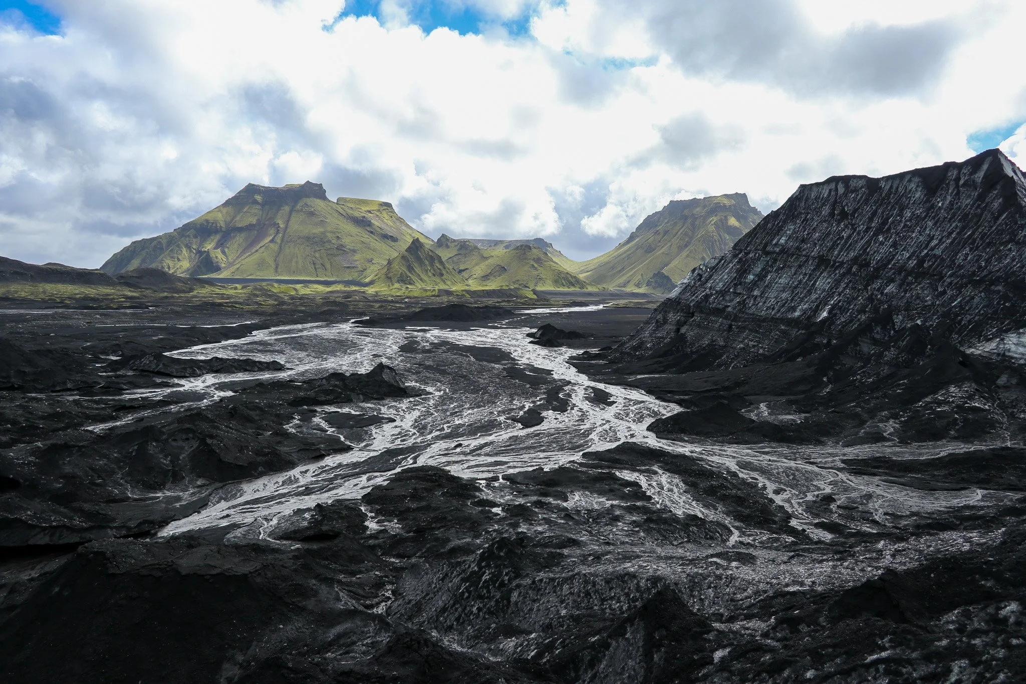 A landscape of volcanic mountains with green moss and black lava rocks, with a river flowing through the dark volcanic terrain under cloudy skies.