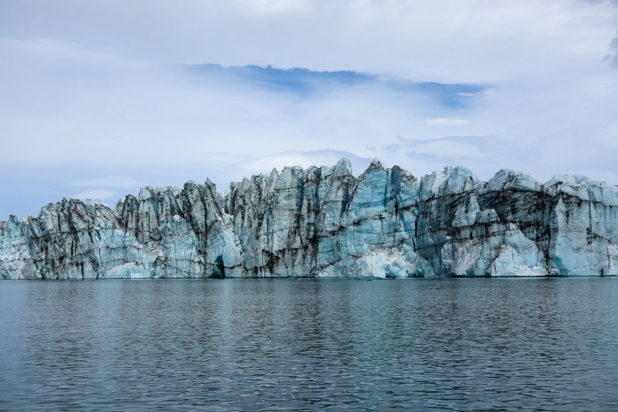 A glacier with jagged ice formations and black streaks of dirt, floating in a calm body of water with a cloudy sky overhead.