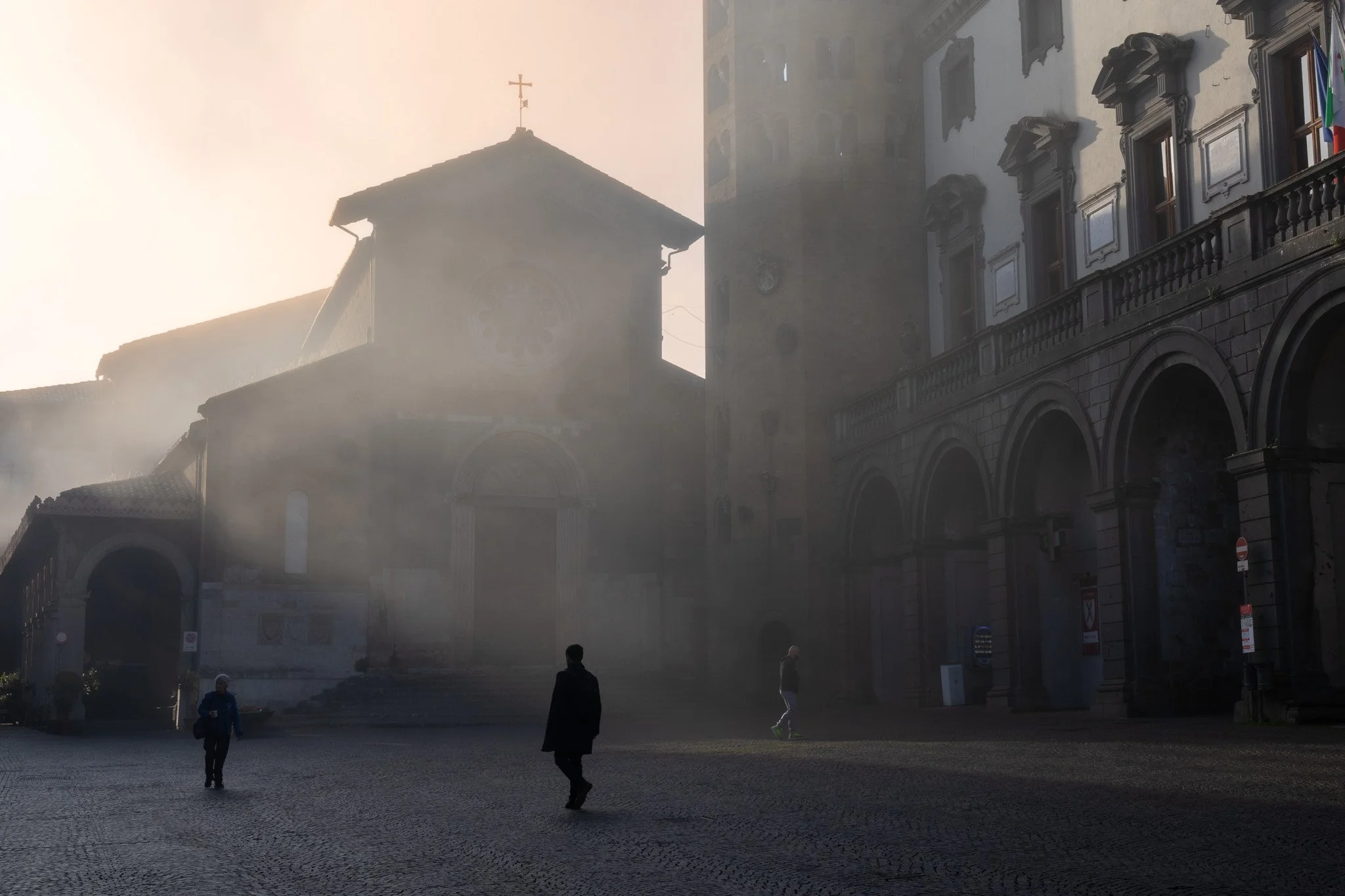 A foggy city square with three people walking, old stone buildings, and a church with a cross on top in the background.