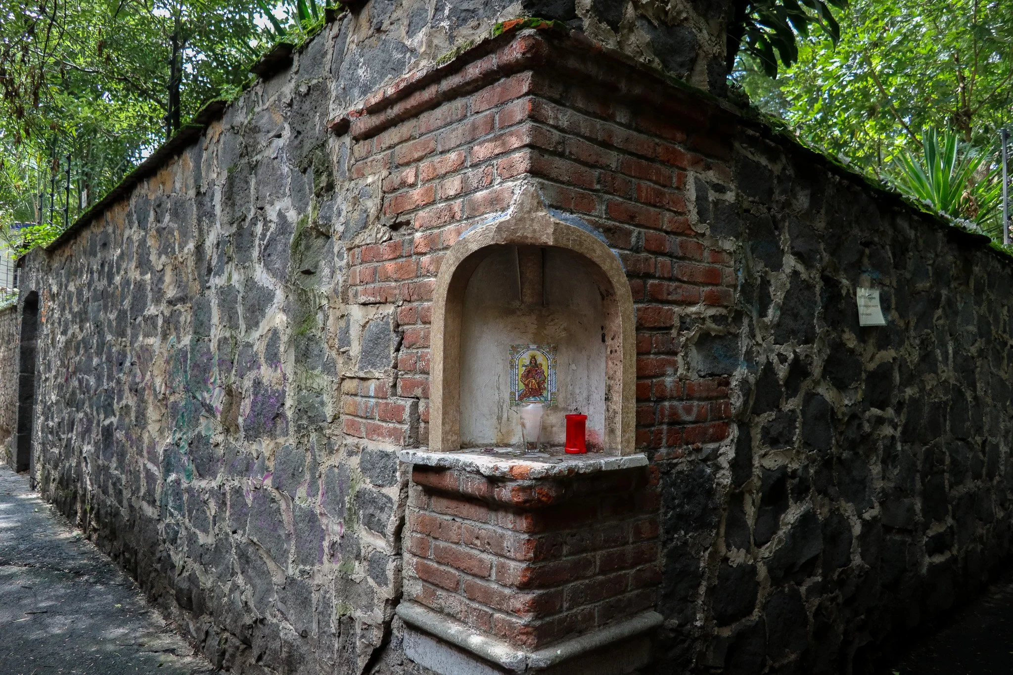 Small religious shrine built into a corner of a stone and brick wall, containing a picture of a deity, with candles and a red candle holder in front of it, surrounded by green foliage.