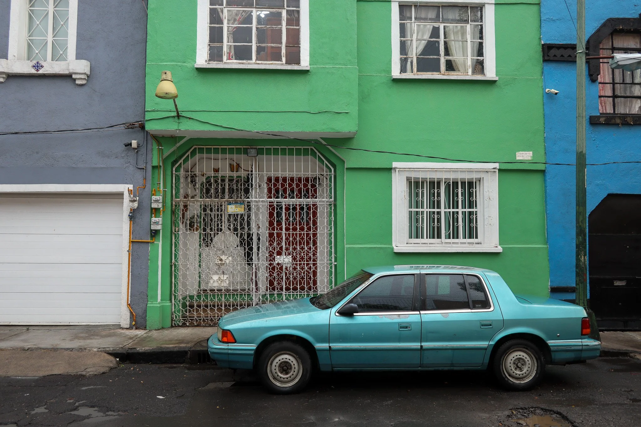 A blue car parked in front of a colorful building with a green and blue exterior walls, white grilles on windows, and a metal gate at the entrance.